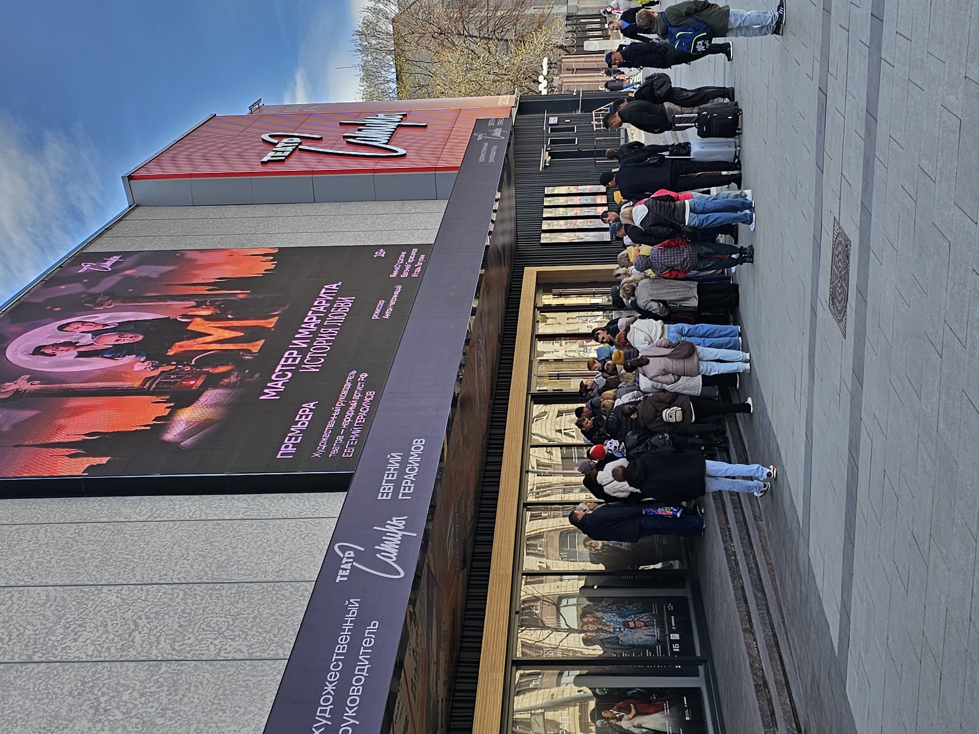 Here is a detailed description of the image:
**Scene Overview:** The image captures a queue of people waiting outside the "Satire Theatre" in Moscow, Russia. The theatre's facade is prominent, featuring large posters and signage.
**Building Details:** The theater has a modern exterior with gray paneling and a red striped top section. Large posters advertise the show "Master and Margarita. A Love Story." Signage identifies the theater as the "Satire Theatre" and lists Evgeniy Gerasimov as the artistic director.
**People:** A long line of people are waiting in front of the theater. They are dressed in everyday winter clothing, including coats, jackets, jeans, and hats. Many are looking toward the entrance of the theatre. Several have suitcases.
**Signage and Posters:** The posters on the theater advertise the play "Master and Margarita," with a promotional image featuring actors. The play is marked as 16+.
**Environment:** The scene takes place outdoors. The sky is blue with some clouds. There are trees visible in the background, suggesting it's an urban environment. The pavement is made of gray paving stones.
**Location:** The location can be identified as the "Satire Theatre" in Moscow based on the signs and promotional materials.
