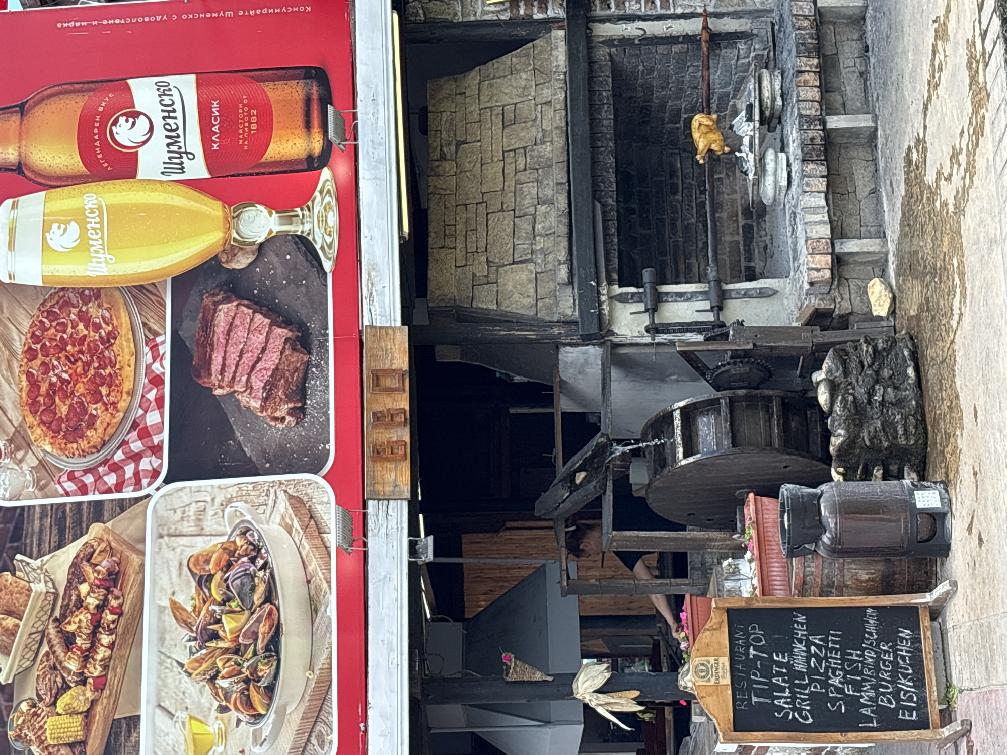 The image shows the exterior of a restaurant with a prominent outdoor grill and a chalkboard menu. The chalkboard lists "RESTAURANT TIP-TOP," followed by menu items such as "SALATE," "GRILL HÄHNCHEN" (grilled chicken), "PIZZA," "SPAGHETTI," "FISH," "LAMM/RIND/SCHWEIN" (lamb/beef/pork), "BURGER," and "EIS/KUCHEN" (ice cream/cake). Behind the chalkboard, a woman is partially visible, appearing to be working near an outdoor kitchen area with a water wheel feature. To the right of the grill, a chicken is roasting on a rotisserie. The overall scene suggests a casual dining establishment, likely in Varna, Bulgaria, given the commonality of such restaurants in tourist areas there. The backdrop includes a large stone structure, likely a fireplace or oven, and various cooking implements. The poster above the restaurant entrance features images of food and drinks, including beer and pizza, consistent with a restaurant setting.
