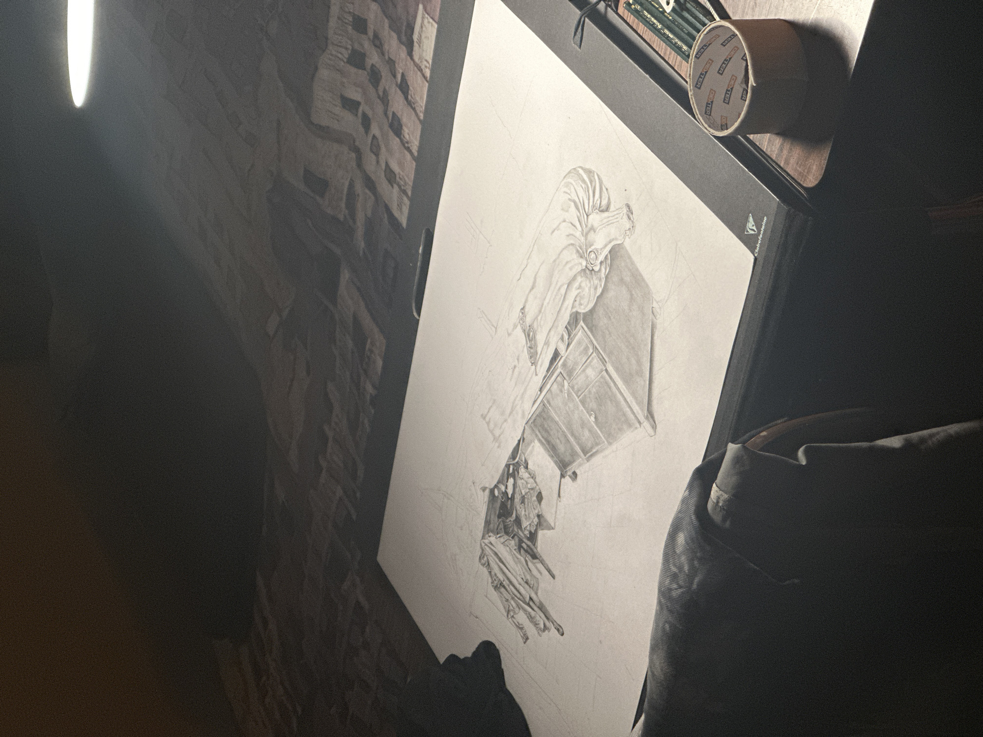 A drawing of a bureau with clothes strewn underneath is on a drawing board. A roll of tape and pencils rest on the board next to the drawing. The drawing is next to a brick wall with a bright light. The location is Montreuil, France.
