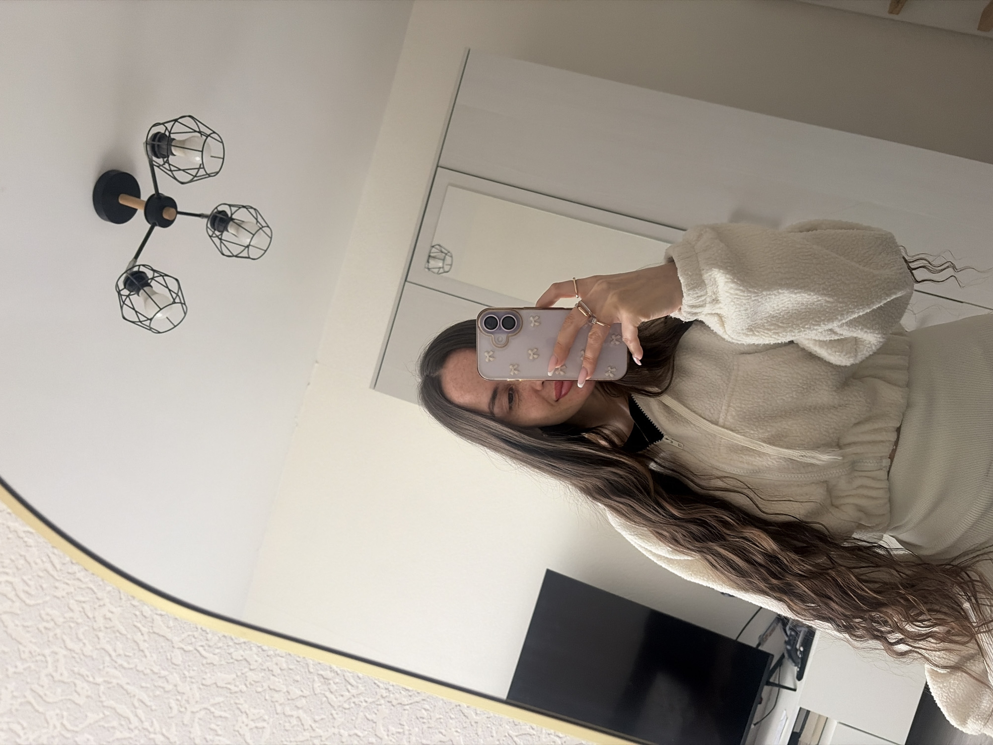 A young woman is taking a selfie in a mirror. The room appears to be a modern living space, with a black television against a white wall and a contemporary chandelier. The woman is wearing a white, cropped sweater and has long, wavy brown hair. She is holding a phone with a floral-designed case. The image does not appear to be taken in Saint Petersburg, Russia.