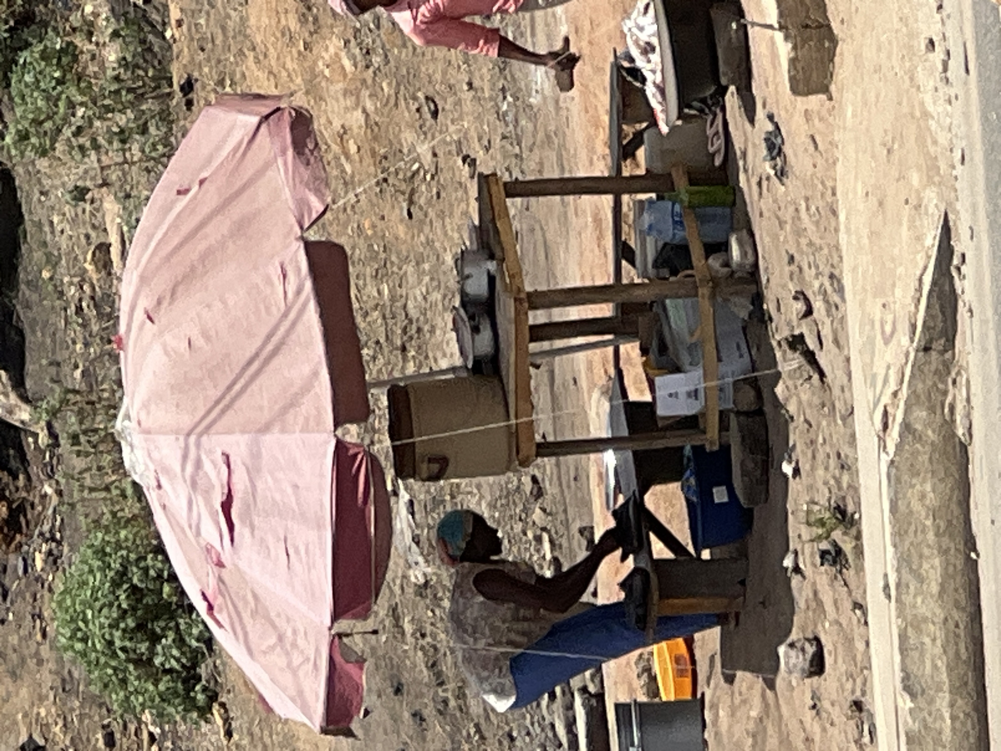 The image depicts an outdoor street scene, likely in Jos, Nigeria, under bright daylight. The main subject is a person seated at a functional, utilitarian wooden stall, actively operating a manual machine that appears to be a sewing machine or similar device. This individual wears a blue head covering, a light-colored patterned sleeveless top, and blue trousers. Their hands are engaged with the machine's mechanism.
The wooden stall is crudely constructed, featuring shelves holding various items like bottles and containers, suggesting it functions as a vending or service point. Providing shade for the stall is a large, light pink umbrella positioned to the left. The umbrella shows signs of wear, with several visible tears in its fabric.
In the upper right portion of the frame, a second person is partially visible, standing in pink clothing. The ground is an uneven mix of dirt, sand, and small rocks, indicative of an unpaved area or roadside. Sparse green vegetation, possibly bushes, grows in the background on the left. To the far right, a lighter, smoother surface suggests the edge of a paved road or sidewalk. The scene conveys a sense of daily life and informal commerce in a sunny, arid environment. No discernible text is visible in the image.