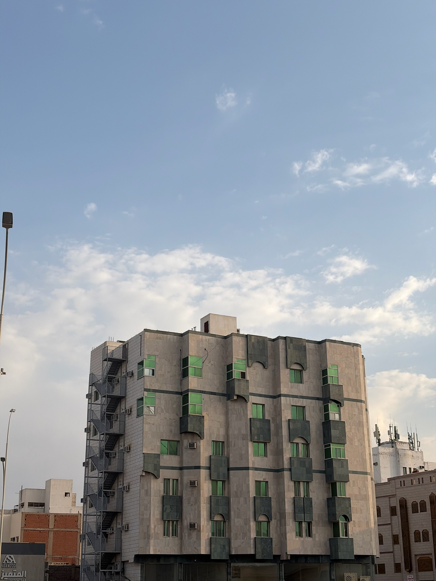 In Medina, Saudi Arabia, a contemporary building with a unique, stepped facade and green windows is captured from a low angle against a sky dotted with white clouds. The building's exterior is clad in light-colored stone, possibly marble, with darker gray stone accents framing the windows and creating decorative vertical elements. An external metal fire escape runs along one side of the building, providing access to multiple floors. Adjacent to this building, other structures are visible, including a white building with antennas on its roof and a red brick building. A street light on a tall pole stands to the left of the main building, and a street-level sign with Arabic text is partially visible at the bottom left. The overall scene suggests an urban environment with modern architecture under a bright, partly cloudy sky.