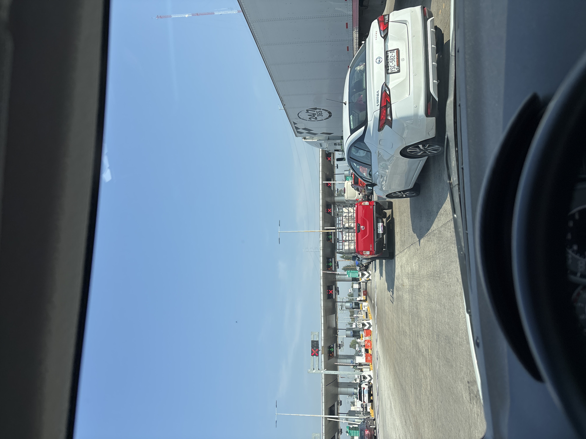 The image shows a view from inside a vehicle, likely a car, at a toll booth. The location is inferred to be near Santa María Moyotzingo, Mexico, based on the license plate of the white Nissan Versa in front (YSC-582-A), which indicates the state of Puebla. Other vehicles are visible, including a large white truck and a red truck. The toll booths are overhead, with lights indicating which lanes are open or closed. The sky is blue and clear. The steering wheel of the photographer's vehicle is visible in the foreground. The scene depicts a typical traffic queue at a toll plaza on a sunny day. There are no visible people or interactions.