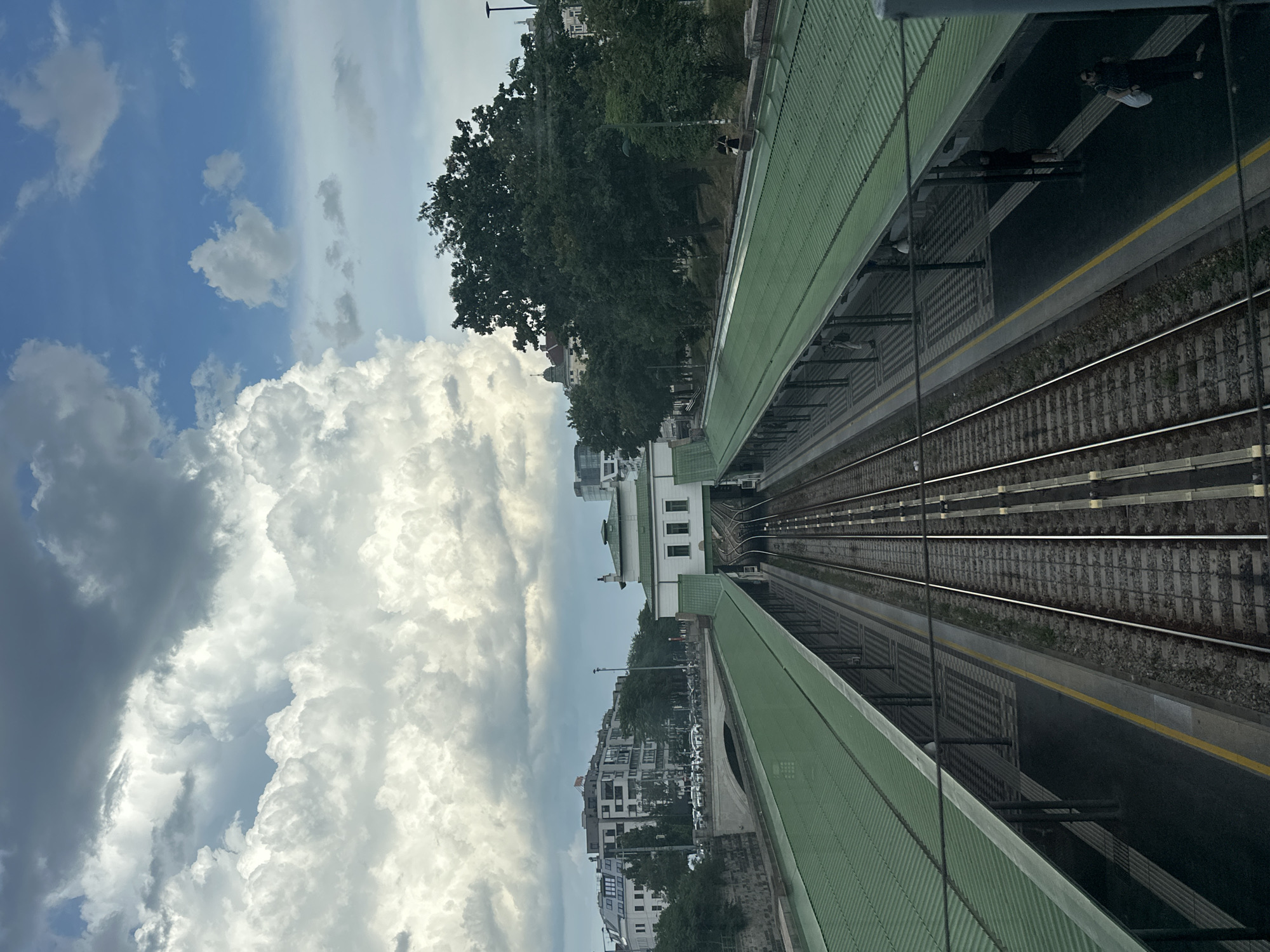 The image captures a view of a train station in Vienna, Austria, likely from an elevated position. The foreground features two sets of train tracks, a typical element of any railway station. The tracks are flanked by platforms covered by striking green metal roofs, supported by dark poles. These platforms are mostly empty, but a few individuals are visible standing on them, some appearing to wait or walk.
Beyond the platforms, the scene transitions to a more urban landscape. A white building with a green roof, possibly part of the station's architecture, is situated in the middle ground. Further in the distance, the cityscape of Vienna unfolds, with several multi-story buildings visible. These structures exhibit a mix of modern and classical architectural styles, characteristic of the city. Trees, both within the station grounds and in the surrounding urban area, add a touch of greenery to the scene.
The sky above is a prominent feature, filled with dramatic clouds. A significant portion of the sky is occupied by large, fluffy white clouds, some of which are illuminated by sunlight, suggesting a bright but potentially dynamic weather day. Patches of blue sky are also visible, indicating a partly cloudy sky. The overall atmosphere is one of a functional urban transit hub set against a backdrop of Viennese architecture and a lively sky.