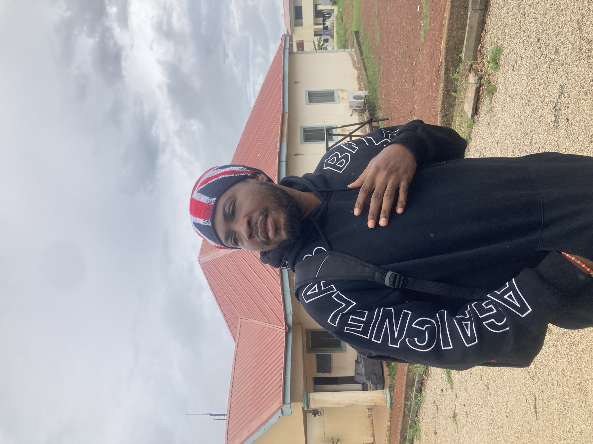 The image, oriented correctly, displays a young man of color posing outdoors in what appears to be Bisichi, Nigeria. He is positioned slightly off-center to the left, looking directly at the camera with a subtle, friendly smile. He has a short beard and mustache.
The man is dressed in a black hooded sweatshirt (hoodie) featuring prominent white block letters on both sleeves, clearly spelling out "BALENCIAGA" multiple times. Over his head, he wears a knitted beanie adorned with red, white, and blue stripes, resembling the design of the Union Jack flag. A black strap from a backpack is visible over his left shoulder, and his right hand is casually placed over his chest.
Behind him stands a building with a distinctive reddish-brown corrugated metal roof and cream or light yellow-colored walls. Several dark-framed windows are visible on the building's facade, along with an outdoor air conditioning unit. The ground in the foreground consists of a mix of gravel and reddish-brown earth, with patches of sparse green vegetation. Another building can be partially seen in the distance behind the main structure.
The sky is overcast with soft grey clouds, indicating a cloudy daytime setting. The overall atmosphere is casual and relaxed.