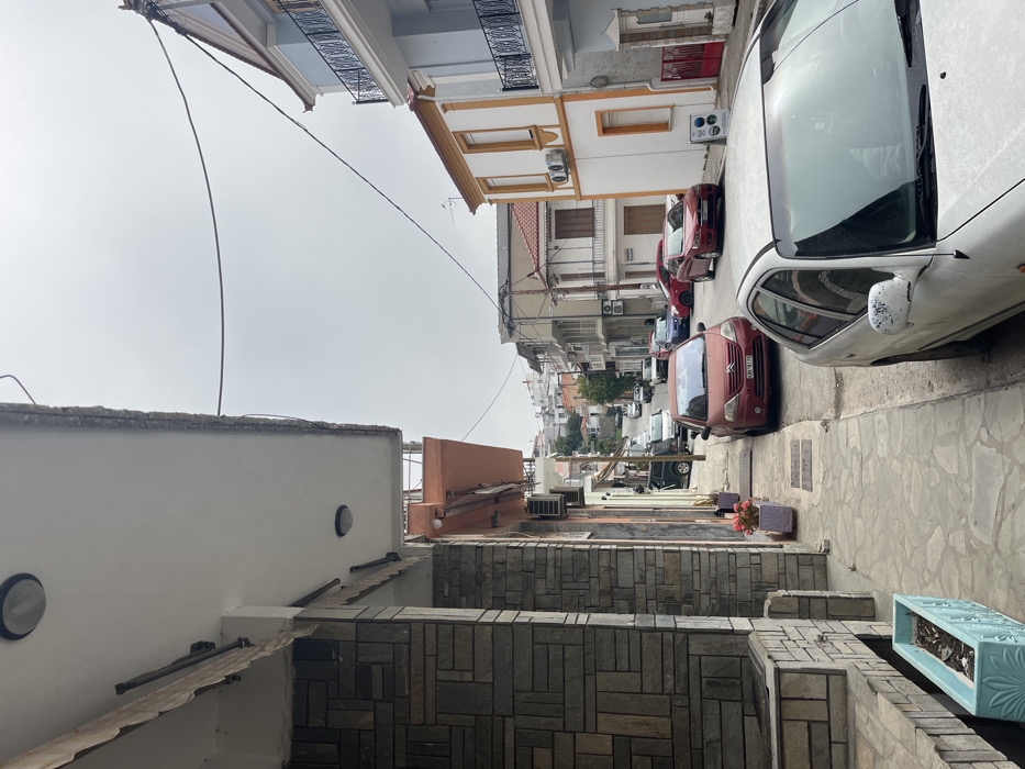 The image depicts a street scene in Limenaria, Greece, located at latitude 40.627 and longitude 24.575. The view is from the sidewalk, looking down a narrow street lined with buildings on both sides. The buildings are primarily two-story, with white stucco walls and terracotta tile roofs. The street surface is paved with gray stone. There are multiple parked cars lining the street, including a red sedan, a white sedan, and a silver hatchback. In the foreground, a person is seated in the driver's seat of a white sedan, looking out the front windshield. The sky is overcast with a pale gray color. The scene is quiet, with no visible people or activity beyond the driver in the foreground.