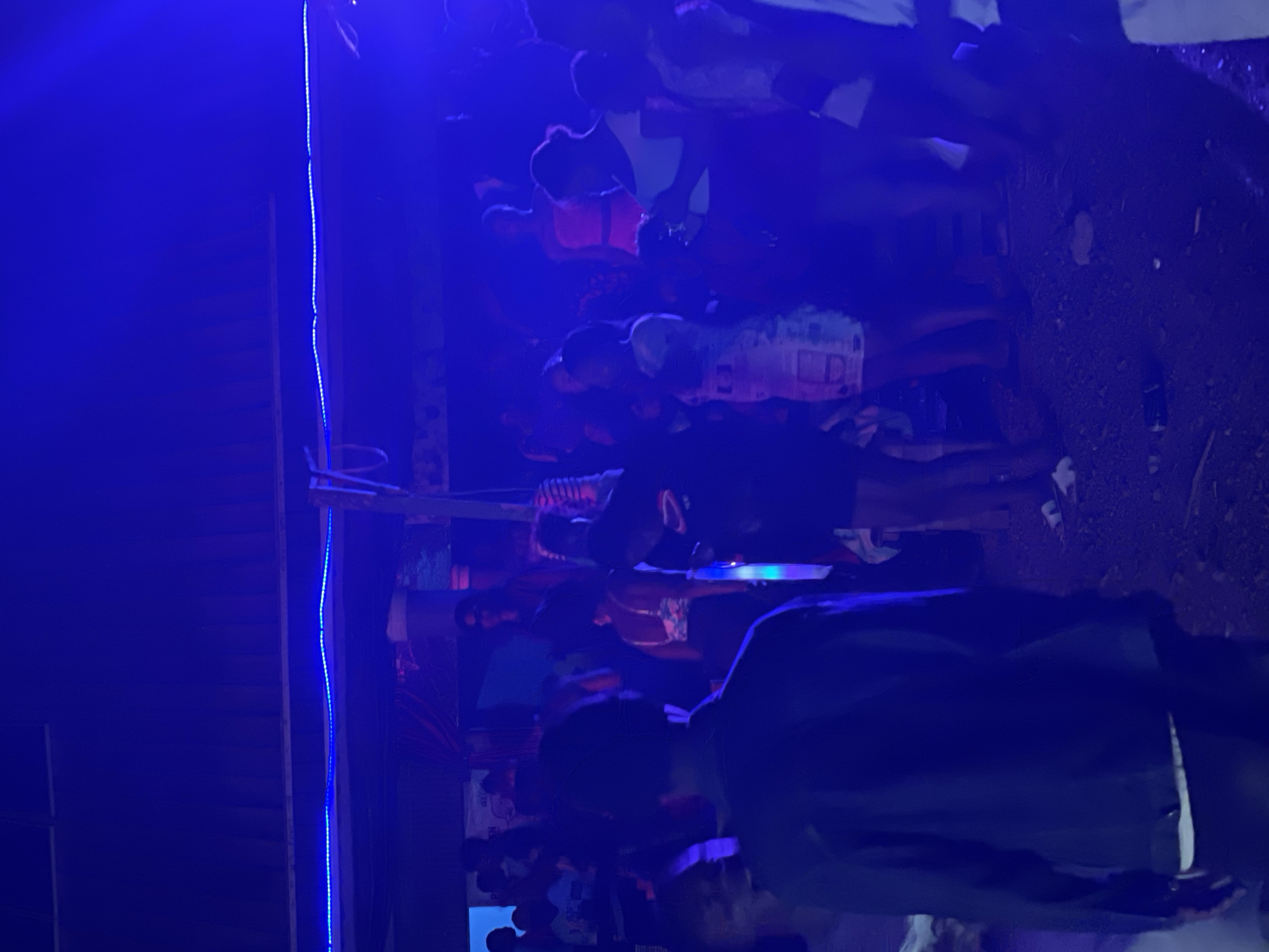This image captures a gathering of people at night, illuminated by blue lighting, suggesting a party or social event. The setting appears to be an outdoor or semi-outdoor space with a covered roof supported by posts. A prominent feature is a string of blue LED lights running along the edge of the roof structure.
In the foreground, a person wearing a dark jacket is seen from the back, facing away from the camera. Behind them, a crowd of individuals is visible, with several people dancing or mingling. Some women are dressed in brightly colored or patterned attire. The ground is uneven and appears to be dirt or gravel, with some scattered debris.
The overall atmosphere is dynamic and energetic, characteristic of a nighttime social function. The blue lighting creates a vibrant and somewhat mysterious ambiance. While the specific location within the city of Durimi, Nigeria, is not explicitly indicated by any visible landmarks or signage, the image depicts a scene that could be found in various community spaces or entertainment venues within the city.