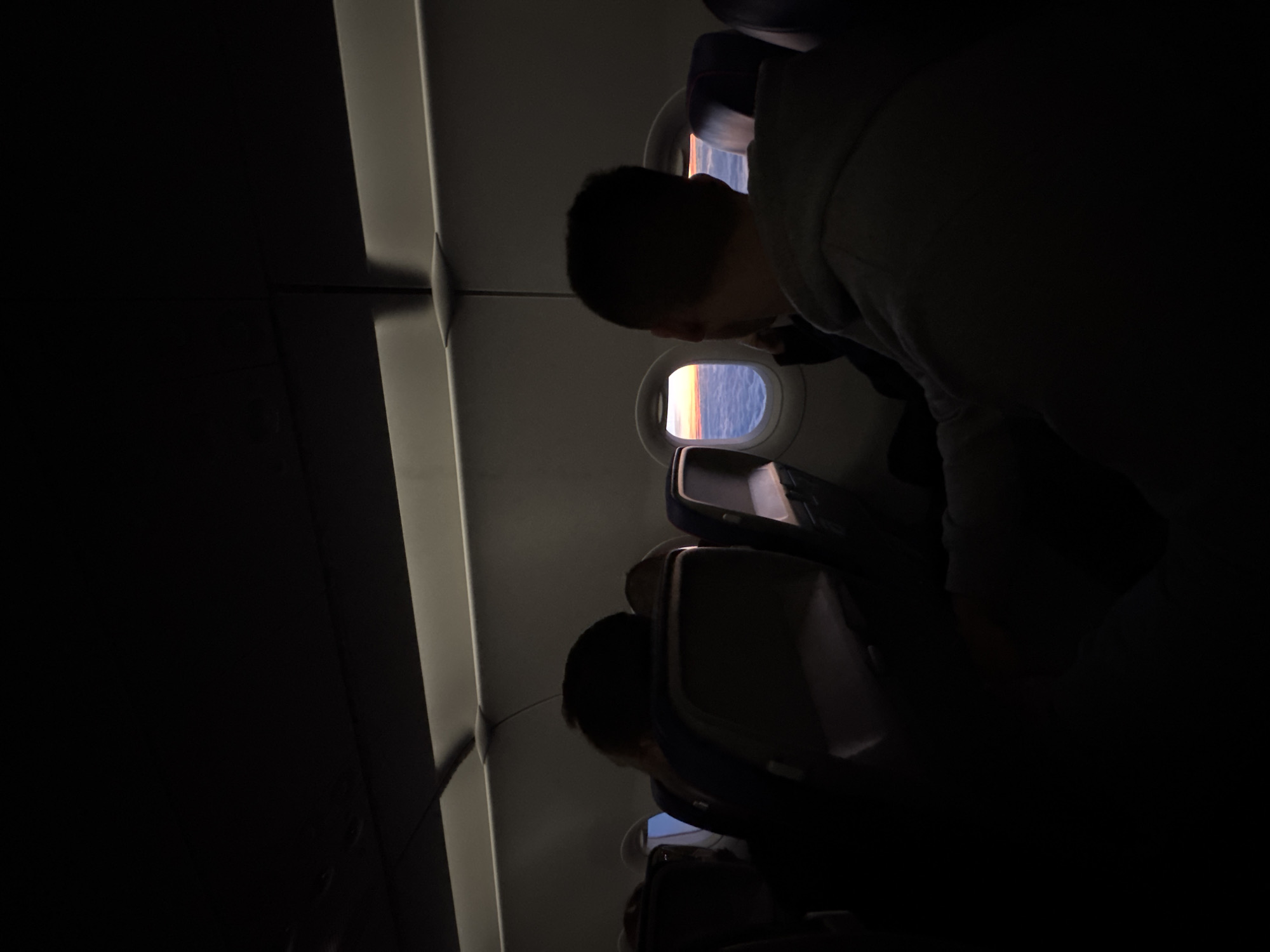 The image shows the interior of an airplane cabin. A passenger is looking out the window. The scene appears to be taken during dusk or dawn, judging from the view outside the window, which suggests a sunset or sunrise over the ocean. The location is not Dortmund, Germany.