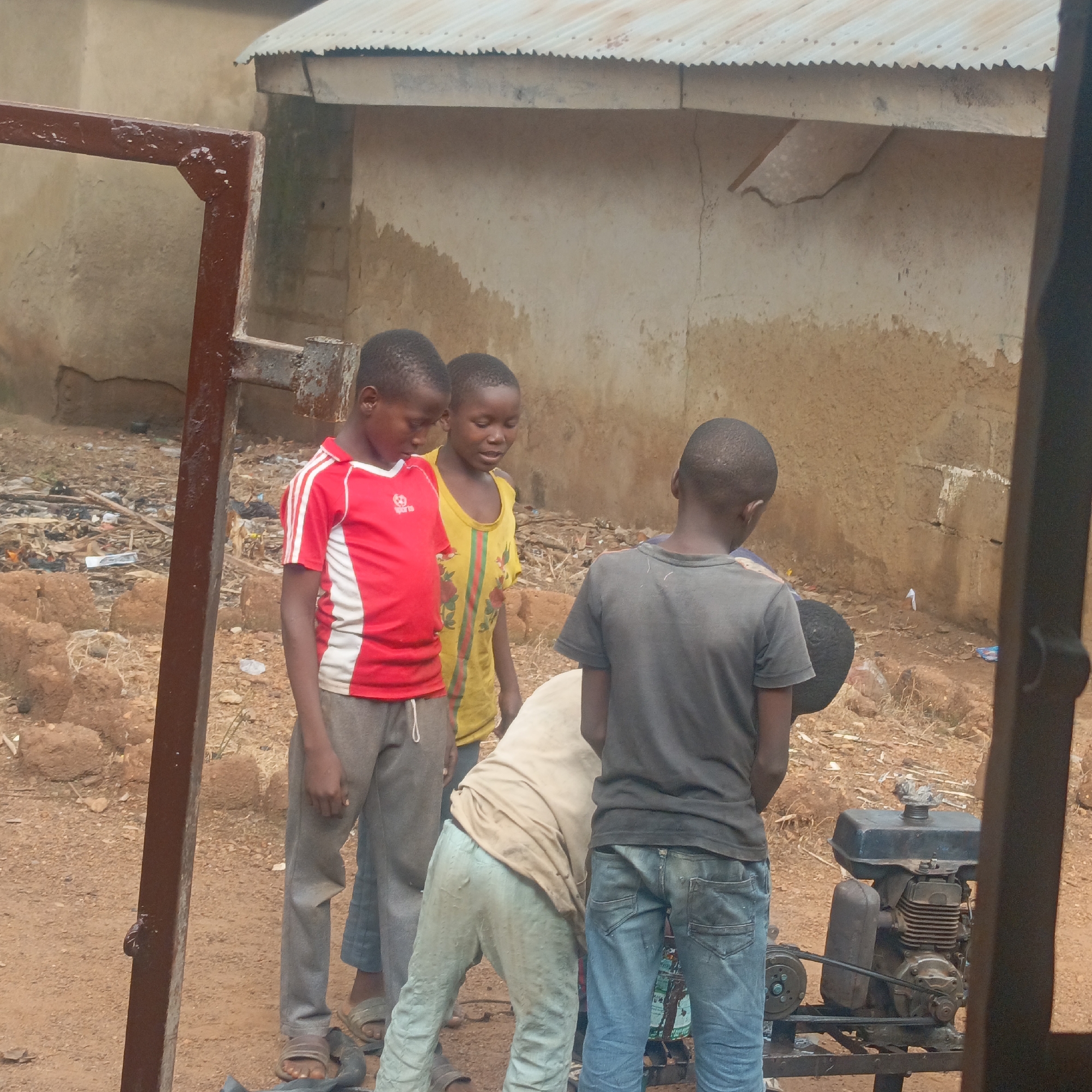 The image depicts a group of young boys gathered around a small engine in what appears to be an outdoor, informal setting, possibly a residential area or a workshop on the outskirts of Akwanga, Nigeria. The background shows a dilapidated, mud-brick structure with a corrugated metal roof, characteristic of many buildings in the region. The ground is dirt and littered with debris, including plastic waste and discarded items, suggesting an area with limited sanitation.
In the foreground, four boys are visible. One boy, wearing a red and white striped shirt and gray pants, stands to the left, looking down. To his right, another boy in a yellow t-shirt with colorful patterns is looking towards the engine. A third boy, facing away from the camera in a dark gray t-shirt and blue jeans, is positioned closest to the engine, seemingly working on it. A fourth boy, wearing a light-colored shirt and light blue pants, is bent over, also appearing to interact with the engine. The engine itself is a small, gasoline-powered unit, likely used for mechanical purposes, possibly for a generator or a small piece of machinery. It is mounted on a metal frame. The overall impression is one of resourcefulness and activity, with the boys engaged in an unknown task involving the engine within their community.