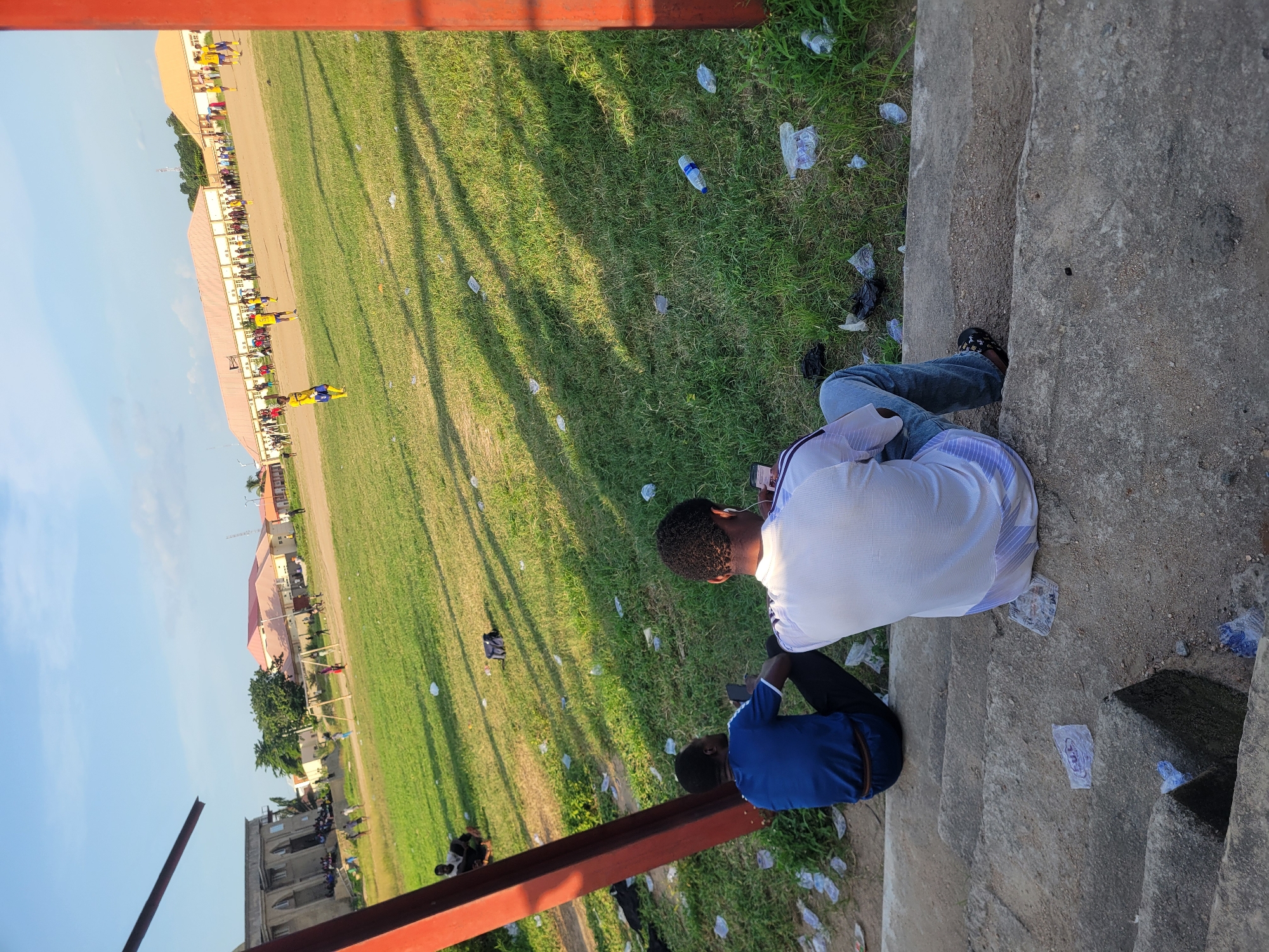 In Lokoja, Nigeria, two men are seated on concrete bleachers, facing away from the camera, looking towards a soccer field. The man on the left wears a blue t-shirt and dark pants, while the man on the right is in a white t-shirt and blue jeans. Both men appear to be using their phones. The field is covered in green grass and is littered with various plastic bottles and bags. Several individuals, some in yellow and blue soccer uniforms, are visible on the field, suggesting a game or practice is in progress. In the background, several buildings, including what appears to be a large, tan-colored structure with a red roof, are visible. The sky is blue with some clouds. A red metal structure is partially visible in the foreground, to the left and right of the frame.