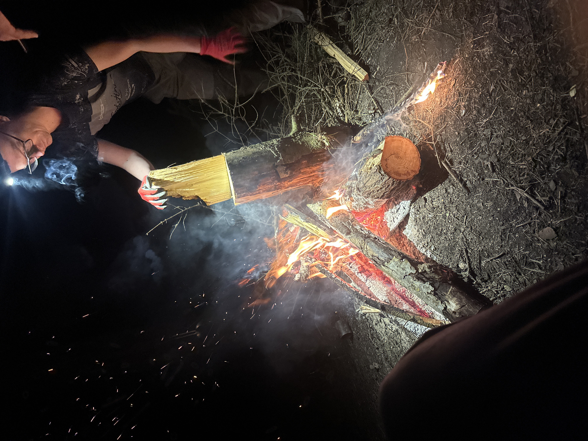 A nighttime scene shows a person with glasses and a headlamp holding a log with red gloves above a campfire. Another person wearing red gloves is partially visible in the upper left of the frame. The campfire is burning with flames and smoke, with sparks flying. There is dry brush and other logs nearby. The location appears to be outdoors and rural. No evidence supports this being within a city, rather the image appears to be in the countryside or wilderness. No city is visible. The image suggests Юзуфоўскі сельскі Савет due to the camera pointing there when the image was taken.