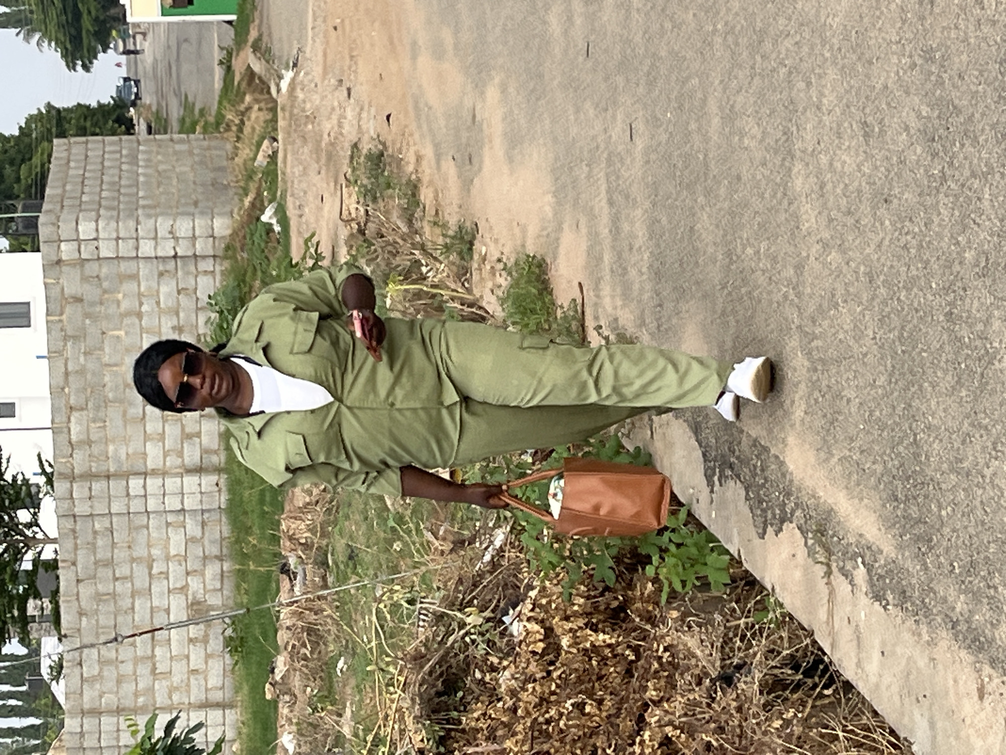A woman in a green uniform, white shirt, and sunglasses stands on a paved surface next to a curb, holding a brown handbag. She also appears to be holding a phone in her left hand. Behind her, there's a concrete block wall and vegetation. The background shows a street with a vehicle and trees. The location appears to be in an urban area, specifically Abuja, Nigeria.
