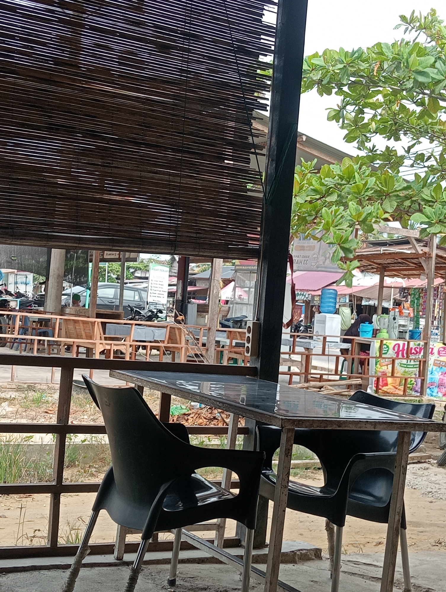 The image depicts an outdoor seating area of a business, likely a restaurant or cafe, located in Lhokseumawe, Indonesia. In the foreground, a black plastic chair is partially visible, placed next to a table with a dark, reflective surface. The table and chairs are supported by a metal frame.
Behind the foreground seating, a wooden railing separates the seating area from a pathway or open space. Beyond this, several other tables and chairs made of wood are arranged. In the background, a row of stalls or shops line a street.
Various objects are visible within the shops, including a blue water dispenser, a blender, and colorful displays of products, some of which appear to be beverages or snacks. A motorcycle is parked near one of the stalls. A sign with text in Indonesian, "PILAT TERAP", is prominently displayed.
The environment suggests a warm climate, with a leafy green tree providing some shade. The overall scene is one of a casual, open-air commercial setting.