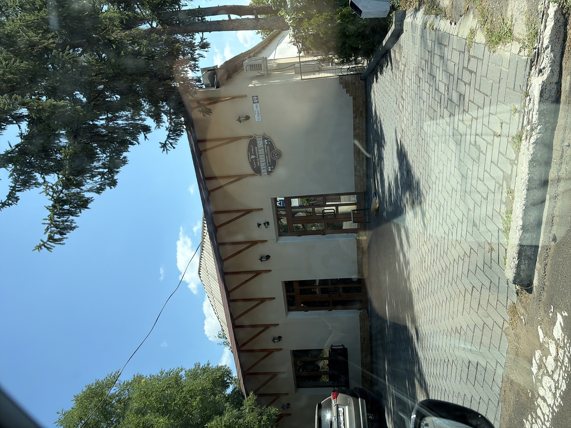 The image shows a building with a light-colored facade, a light-colored tiled roof supported by wooden beams, and a paved driveway in front. A silver SUV is parked to the left of the building. Above the entrance to the building, there is a sign that reads "CRAMA LUCI VASILE" with additional text below. The building appears to be a business, possibly a winery or restaurant, given the sign's content. The area around the building is somewhat overgrown with green trees and bushes. The sky is blue with some scattered clouds. A closer look at the sign reveals text in Romanian, and the address "31 August 1989 49" is also visible. This address, combined with the Romanian text on the sign, suggests the location is in Criuleni, Moldova. The overall scene depicts a sunny day in a semi-rural or suburban setting.
