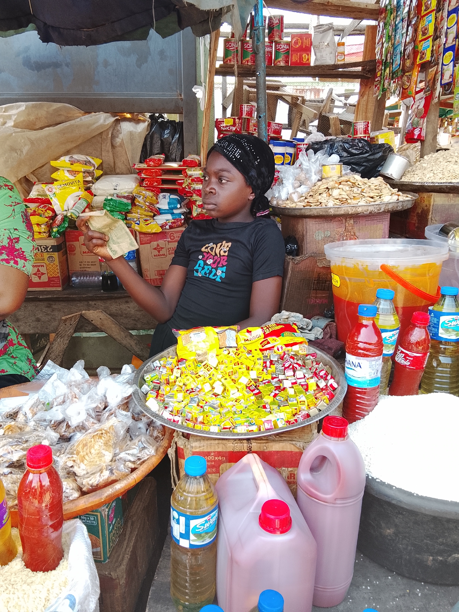 The scene depicts an outdoor market stall located in Akwanga, Nigeria. A young dark-skinned female, likely a vendor, is seated behind a rough wooden counter, looking towards the right. She wears a black t-shirt with the phrase "LOVE YOUR SELF" in multicolored letters and a black headwrap. Her left hand is holding several folded Nigerian Naira banknotes. Partially visible to her left is another individual, whose arm and a green floral-patterned garment are visible.
The stall is laden with various goods for sale. In the immediate foreground, a woven basket contains multiple clear plastic bags of dried goods. To the right, a red-capped bottle with red liquid stands next to a plastic bottle of "Swana" cooking oil. Two large plastic jerry cans filled with red liquid (likely palm oil) and a large black basin containing white grains (likely rice) are also visible.
On the counter directly in front of the vendor, a large metal platter holds a mix of individually wrapped yellow and red seasoning cubes or candies. Behind this, a large clear plastic bucket with an orange handle contains a two-toned substance, yellow over red. To the right of the vendor, another metal platter is piled with dried, light-colored food items, possibly root vegetable chips. Bottles of red liquid and cooking oil are also arranged on the counter.
The background features shelves constructed from wooden planks and metal supports. These shelves are stocked with various packaged goods including canned tomato paste (e.g., "Sonia" brand), bags of processed grain (e.g., "Semovita"), and numerous small packets of spices or seasonings hanging from strings. Cardboard boxes, some branded "Maggi," are also present. An overhead dark fabric canopy provides shade for the stall. The environment suggests a bustling commercial setting.