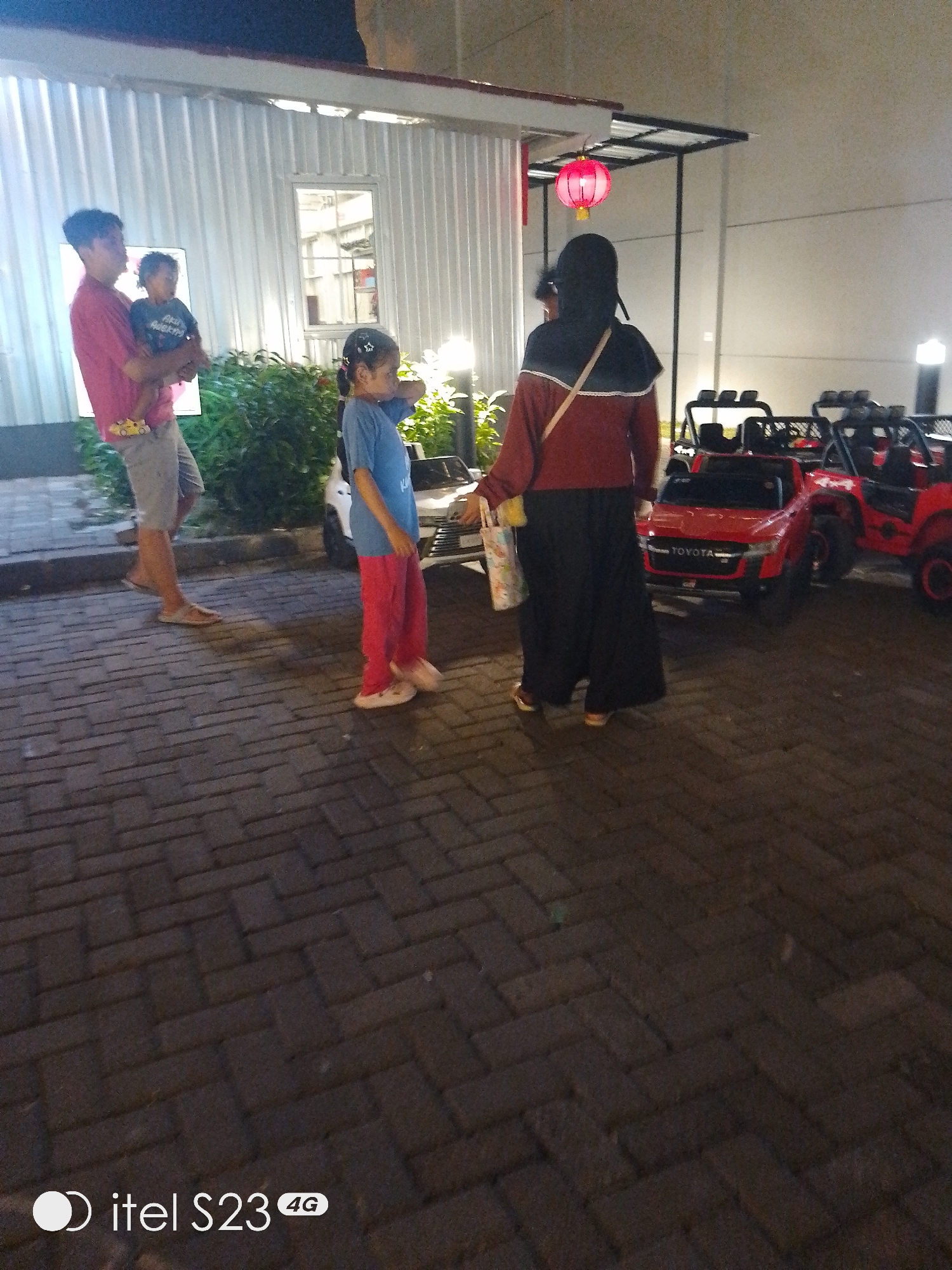 In Surabaya, Indonesia, at night, a man in a pink shirt and grey shorts holds a young child. Beside them, a girl in a blue shirt and pink pants stands, looking away. Behind them, a woman wearing a headscarf and a red and brown jacket walks towards a row of red toy vehicles, some of which are modified to resemble jeeps with roll cages. One of the toy vehicles has "TOYOTA" written on its front. A red lantern hangs from a structure above and to the left of the woman. The ground is paved with brick. The scene appears to be an outdoor area, possibly a parking lot or a recreational space.