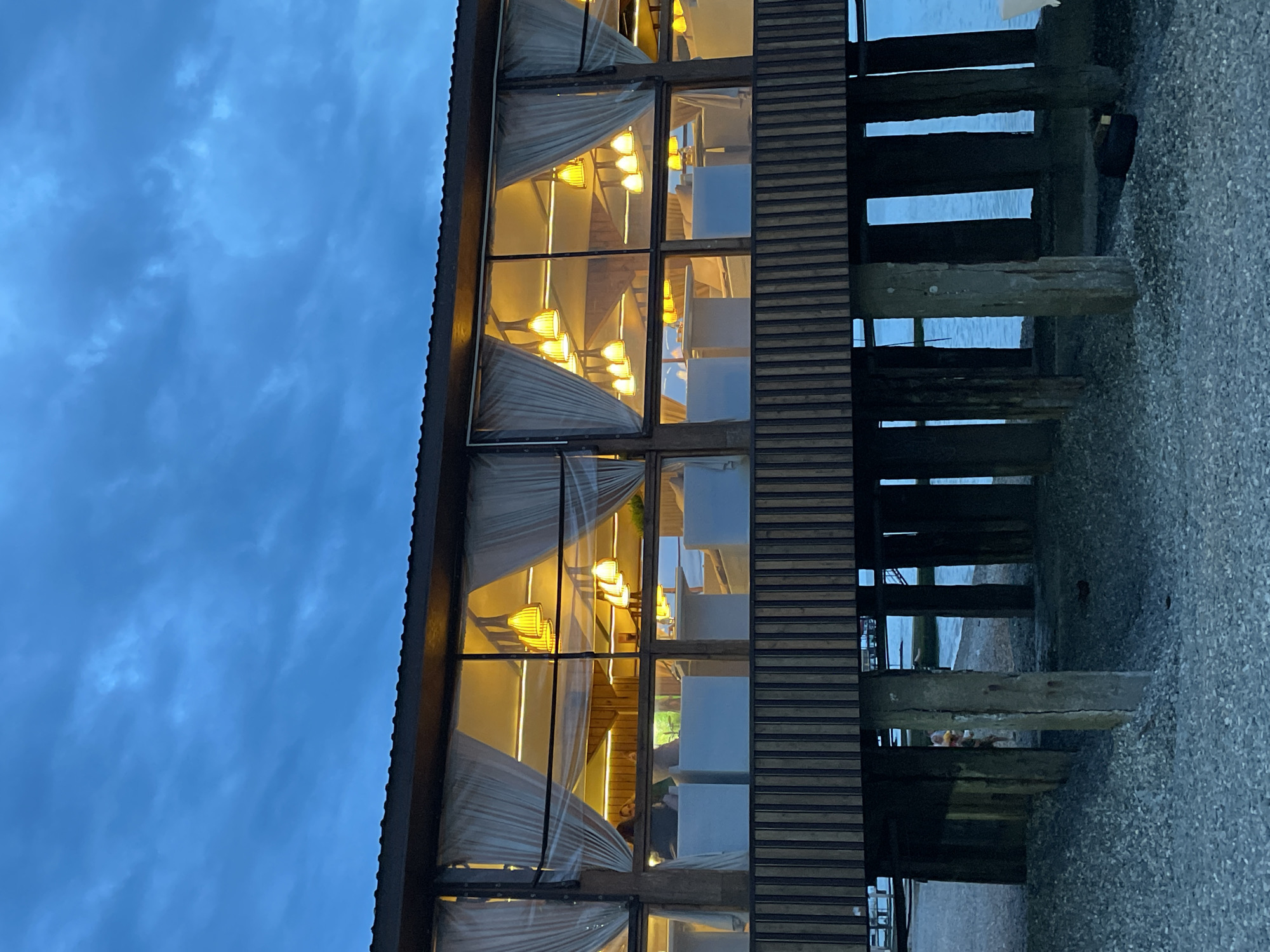 The image shows a building in Sochi, Russia, likely a restaurant built over water. The building features large windows with white curtains partially drawn. Warm light emanates from within, illuminating lamps and suggesting the presence of tables and chairs. The exterior is constructed with dark wood, and the building sits on wooden pilings or posts that extend into the water. The background includes a dark blue sky, possibly indicating twilight. A concrete or pebble-covered ground is visible. The photograph has been taken at an angle.