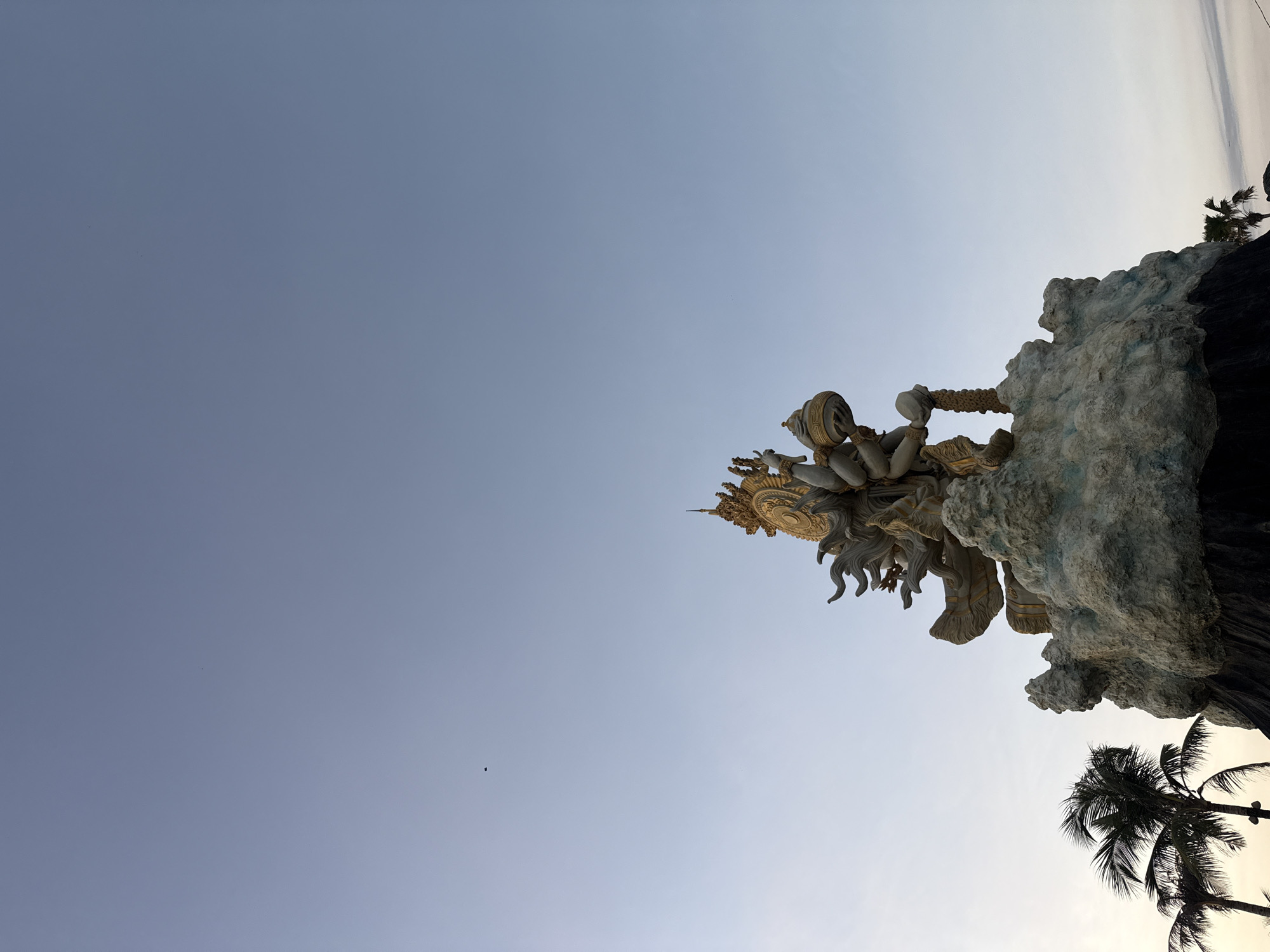The image shows a large, ornate statue with multiple arms, positioned atop a rocky, cloud-like structure. The statue is predominantly gold and gray, with intricate detailing. Palm trees are visible in the lower left and right corners of the frame. The sky is a clear, pale blue. The statue is located in the city of North Kuta, Indonesia. There are no people or animals visible in the image. The scene appears to be outdoors during daylight hours, possibly late afternoon or early morning given the soft light.