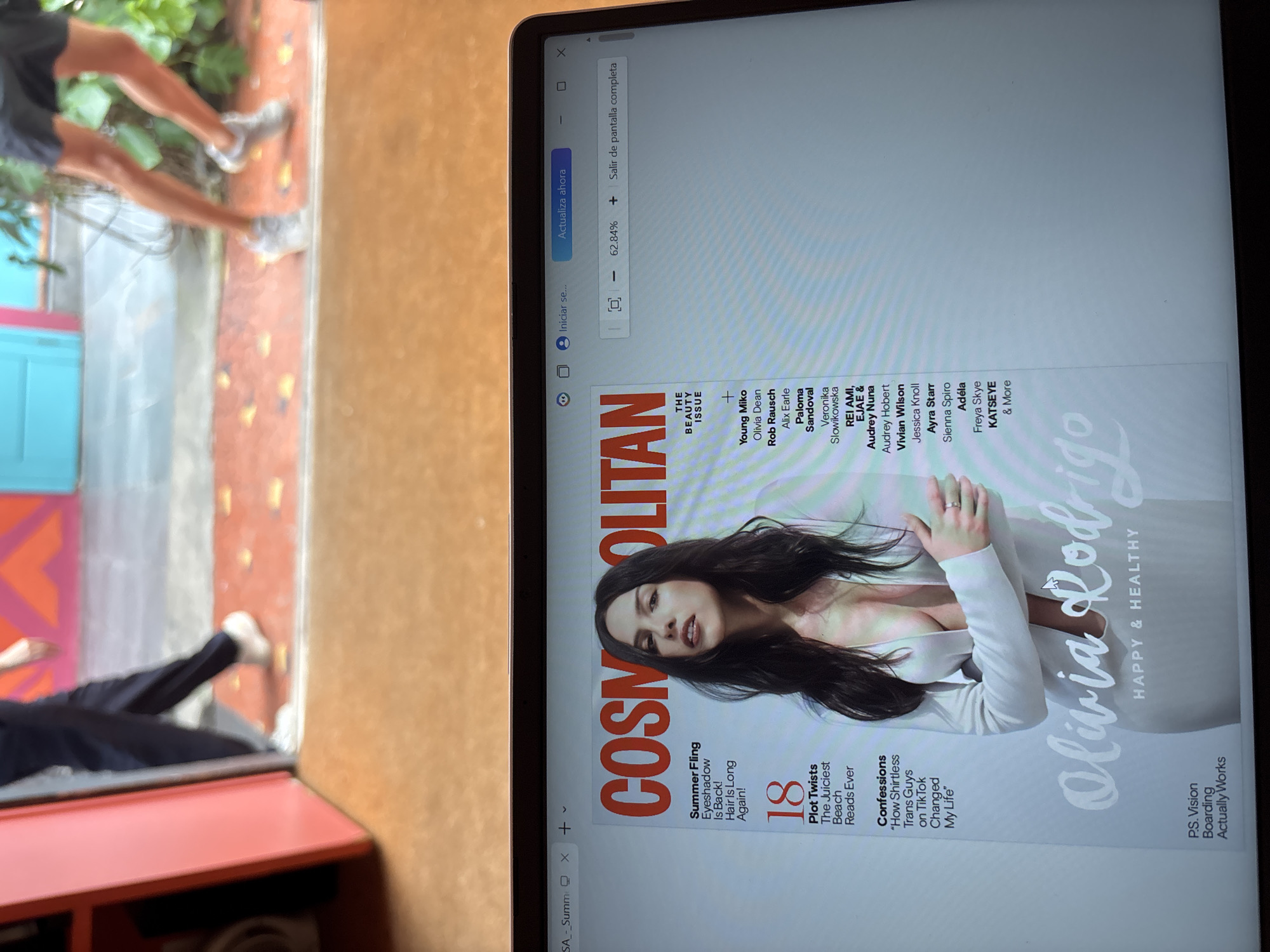 The image shows a laptop screen displaying a digital version of Cosmopolitan magazine with Olivia Rodrigo on the cover. The laptop is resting on a reddish-brown surface. Outside the frame of the laptop, there is a blurred background showing a person’s legs, a colorful wall (red, orange, blue), and greenery. The laptop screen displays a magnified view (62.84%) of the magazine cover, showcasing the Cosmopolitan logo, Olivia Rodrigo's photo, and various article titles. The surrounding environment suggests an indoor setting.