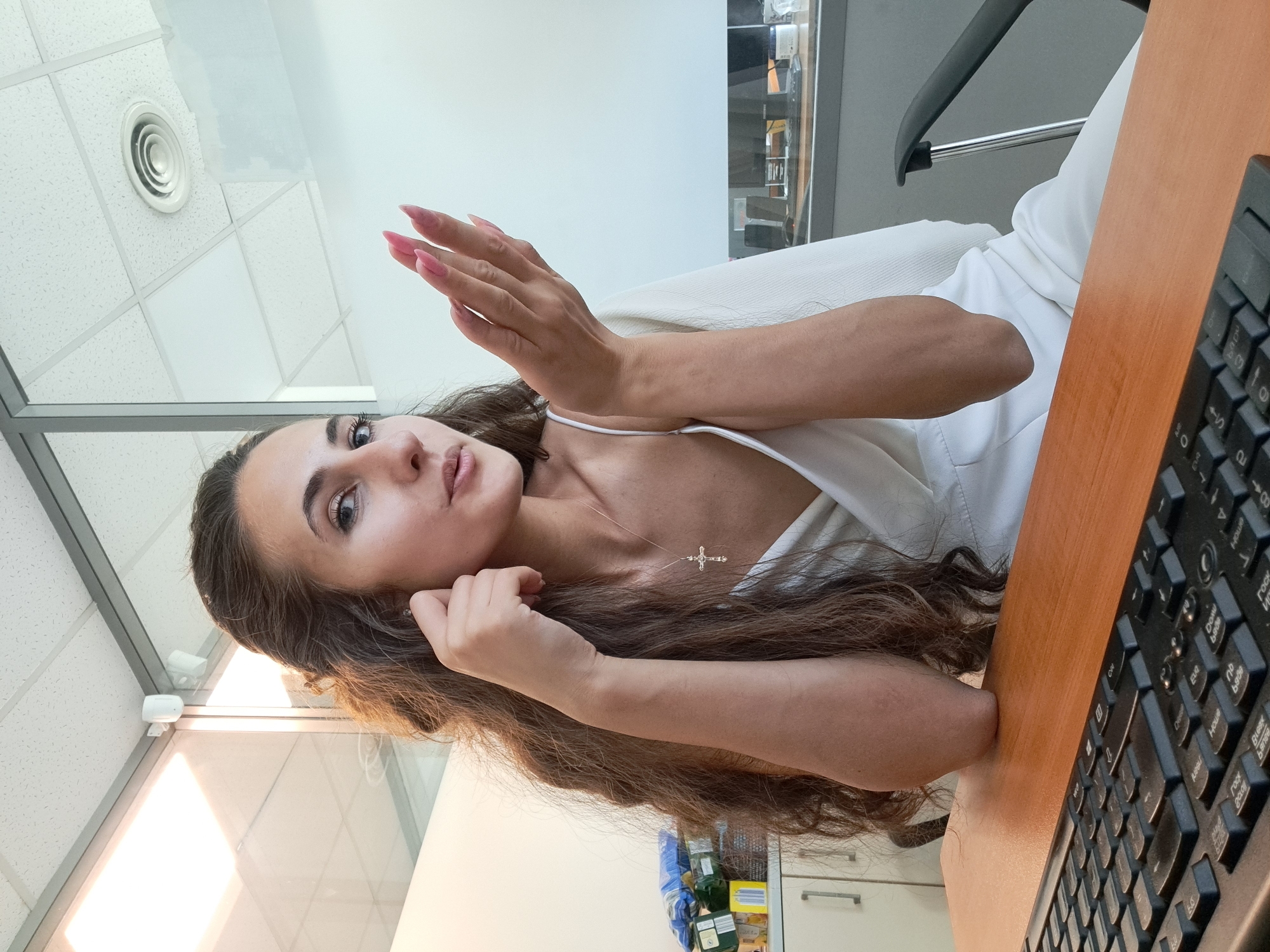 This is a portrait-oriented, close-up, low-angle shot of a young woman indoors, seated at a wooden desk with a black keyboard in the foreground. She has long, dark brown, wavy hair and is wearing a white top, revealing a delicate cross necklace. Her left hand is raised and positioned near her face, with long, pink acrylic nails. Her right arm is resting on the desk.
The background suggests an office or commercial space, with white ceiling tiles, a circular air vent, and large windows or glass partitions. In the distance, shelves with various items are visible, as well as a black office chair. The lighting appears to be artificial, creating a bright, slightly cool atmosphere. The woman's expression is somewhat playful, with her gaze directed towards the camera. No specific time of day or weather conditions can be inferred. The visible text on the keyboard is in Cyrillic.