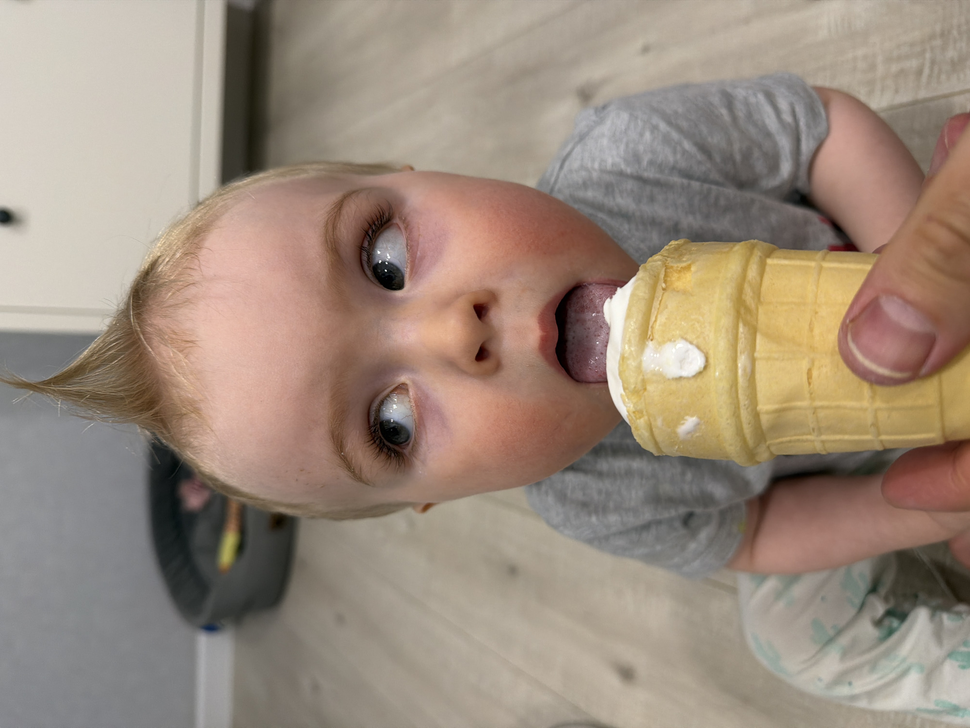 A child is eating an ice cream cone. The child has blonde hair, blue eyes, and is wearing a grey shirt. A hand is holding the ice cream cone up to the child's mouth. The child is licking the ice cream. The background is a light-colored wall and floor. The location of the image cannot be determined as being in Votkinsk, Russia.