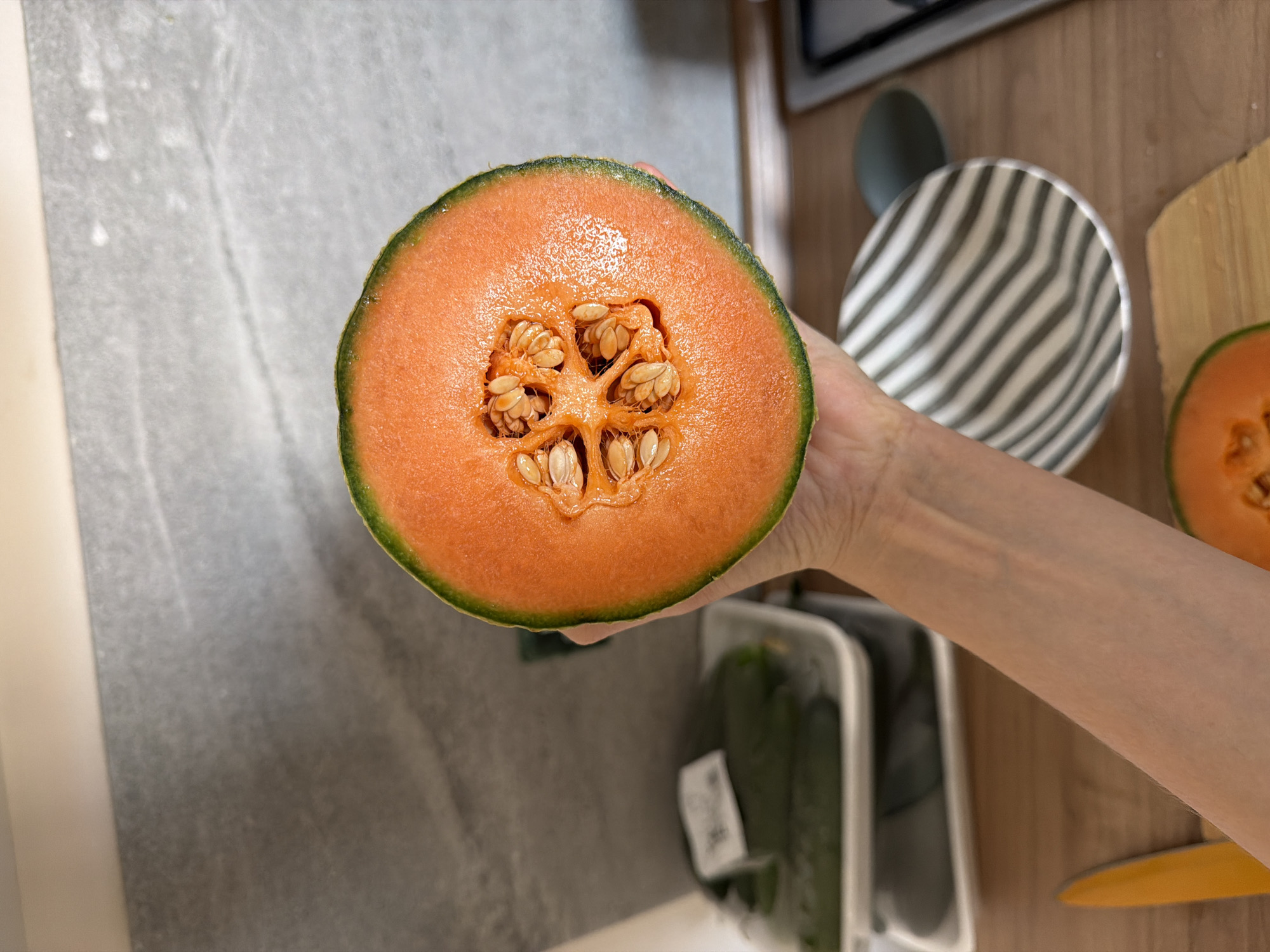 A close-up, high-angle shot depicts a person's hand holding half of a cantaloupe melon, sliced to reveal its orange flesh and seed cavity. The melon has a green rind. The background shows a kitchen setting with a grey countertop, a partially visible white plastic container holding dark green produce, a wooden cutting board with another melon slice, and a grey striped bowl. A knife with a yellow handle is also visible on the wooden surface. The location is suggested to be Catania, Italy, although there are no explicit visual cues confirming this.