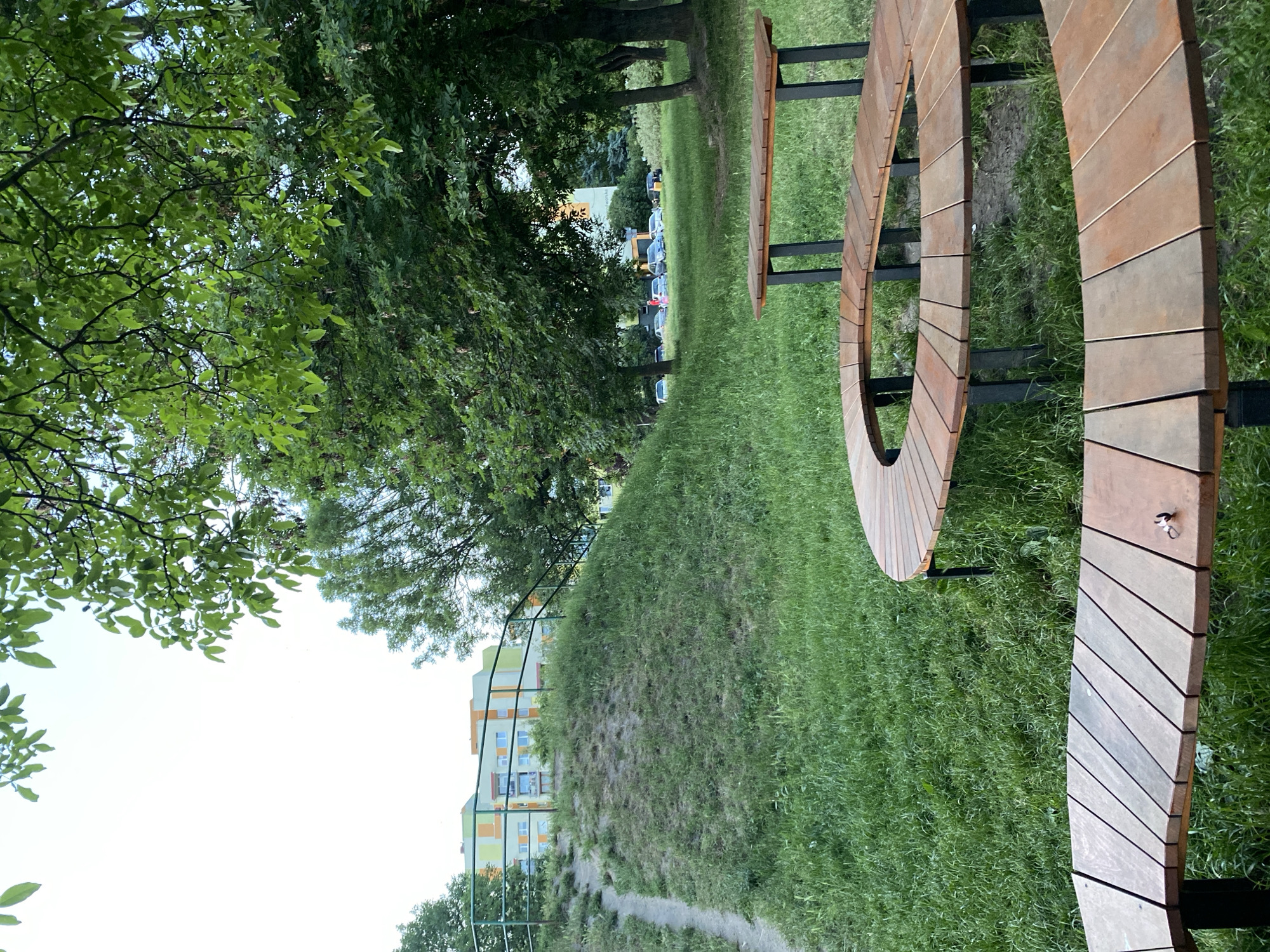 This image depicts a park area in Wrocław, Poland, featuring a gently sloping grassy hill with a worn path winding up its side. A green metal railing runs along the top of the hill. In the background, apartment buildings with colorful facades of green, yellow, and orange are visible. Several cars are parked in front of the buildings. In the foreground, two curved wooden benches with black metal legs are placed on the grass. The bench closer to the viewer has a small silver object, possibly a keychain, on its surface. The scene is set amidst lush green grass and trees, with a bright, overcast sky.
