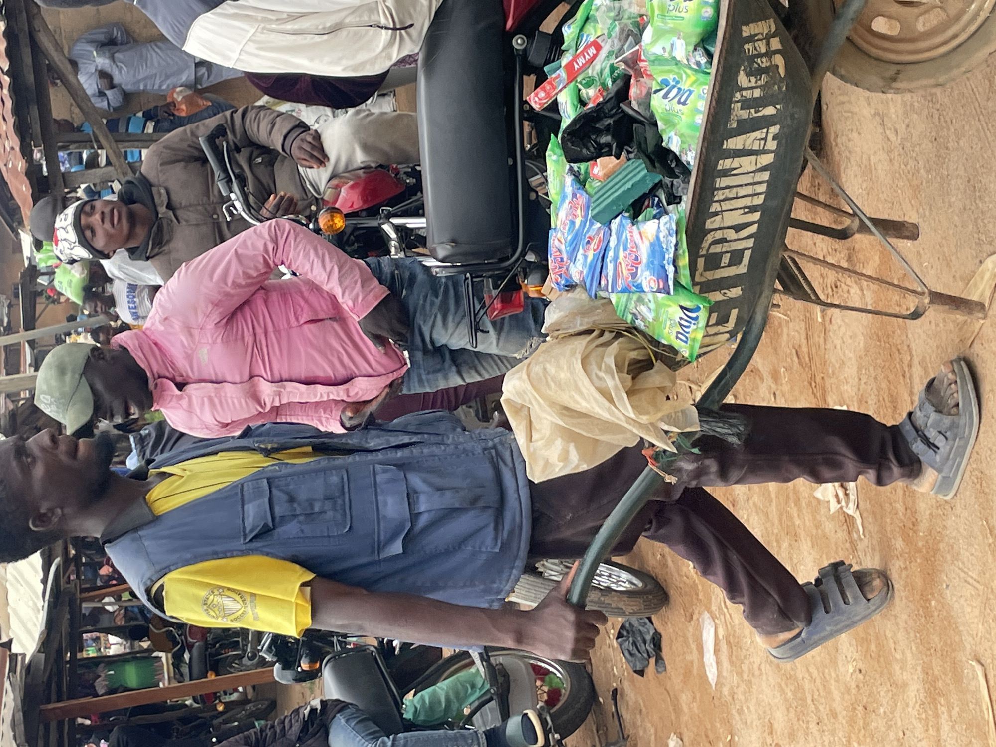 The image depicts an outdoor market scene, likely in the city of Wang, Nigeria. Two men are the primary subjects in the foreground. The man on the left, wearing a yellow t-shirt and a blue vest, is pushing a wheelbarrow filled with goods, including packages of detergent and some loose plastic bags. He is wearing dark pants and sandals. To his right, a man in a pink jacket and blue jeans is walking alongside him. He is wearing a green cap. The wheelbarrow has "DETERMINATION" stenciled on its side. In the background, other people, motorcycles, and market stalls are visible, suggesting a bustling commercial environment. The ground is dirt and sand.