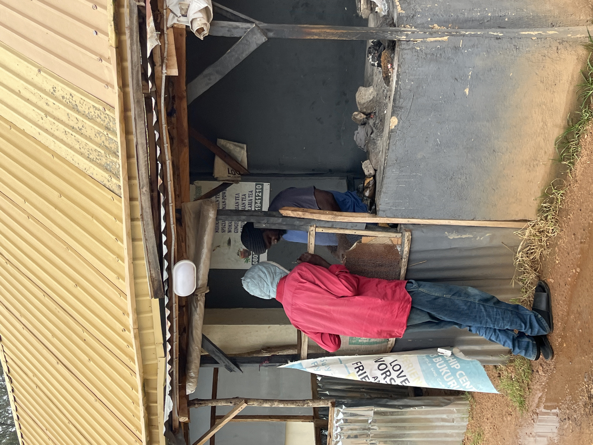 The image depicts an outdoor scene in Bukuru, Nigeria, focusing on a small, open-air stall with a corrugated yellow metal roof and wooden supports. Two individuals are present at the stall. One, wearing a red long-sleeved shirt and blue jeans, stands with their back to the camera, looking into the stall. They are also wearing a light blue head covering and dark sandals. The second individual, wearing a blue t-shirt and a dark beanie, is partially visible inside the stall, interacting with the first person.
The stall appears to be a food or beverage vendor. A sign behind the stall lists various tea products, including "BIAN TEA," "WOMAN TEA," "GINGER TEA," and "LARIA TEA," along with a phone number. The counter area of the stall shows some discarded items and what appear to be cooking implements or fuel. To the left of the stall, a white and blue banner with text including "SHIP CENT," "LOVE," and "VORS" is attached to corrugated metal sheeting. The ground is muddy with puddles, indicating recent rain, and some sparse grass is visible. The overall environment suggests a humble, functional commercial area.