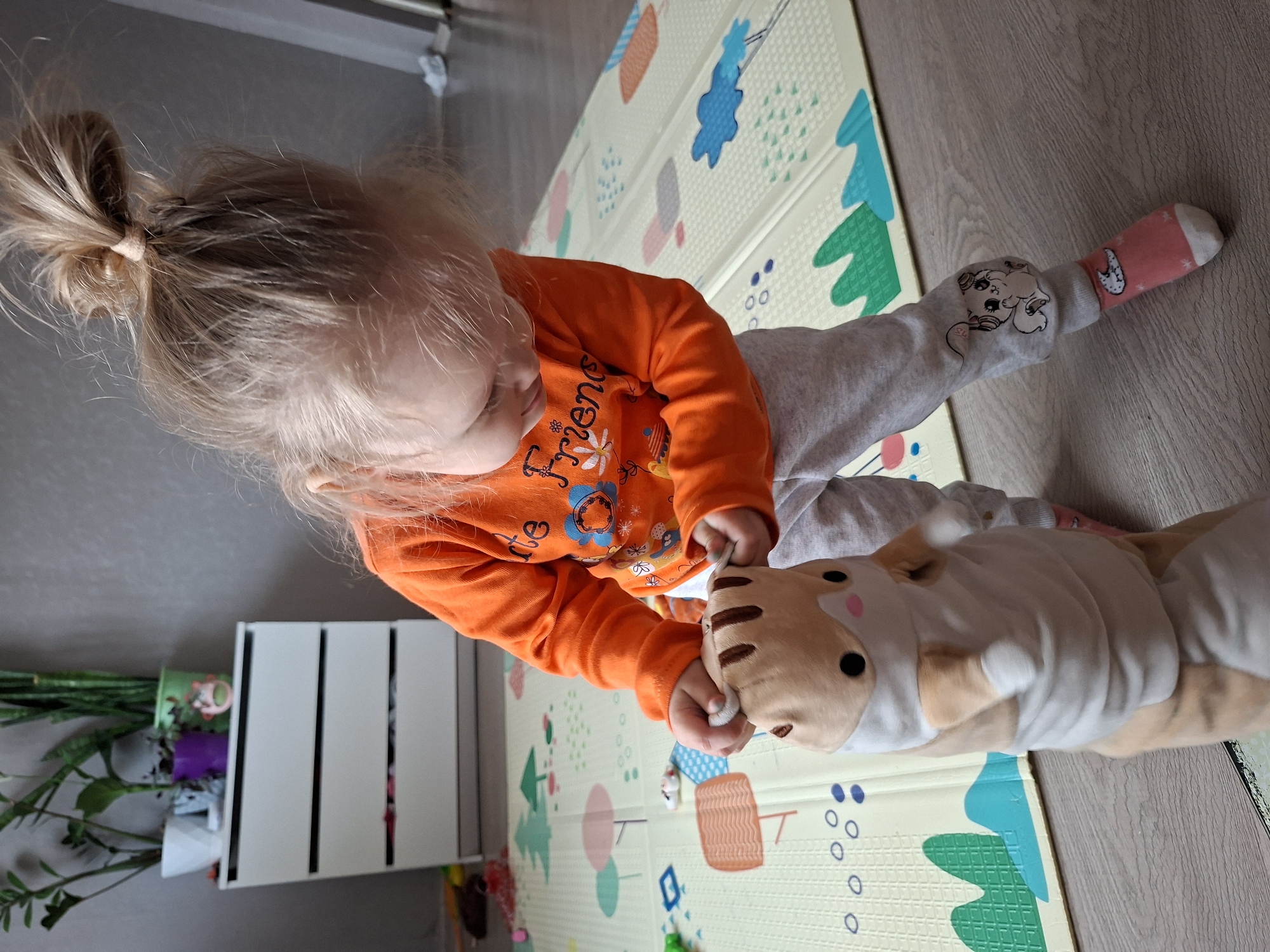 A young child with light-colored hair, tied in a messy high bun, is pictured from an overhead perspective, actively engaged in play on an indoor floor. The child is wearing an orange long-sleeved shirt, grey sweatpants, and a pink and white sock on their visible foot. The orange shirt features white text that reads "FRIENDS" along with a floral design. The grey sweatpants have a graphic of a cartoon character, possibly a dog or cat, on the left leg.
The child is leaning forward, holding the head and neck area of a large, light brown and white striped plush toy, resembling a tiger or similar feline, with both hands. The toy has small black eyes and pink accents on its cheeks. The child appears focused and engrossed in playing with the toy.
They are situated on a light-colored foam playmat with colorful patterns, including abstract trees, circles, and other shapes in blue, green, orange, and pink. The playmat rests on a wooden laminate floor. In the background, to the left, a white slatted organizer or container holds several green plants or artificial flowers. The scene depicts a quiet, playful moment indoors, likely during the daytime given the natural lighting.