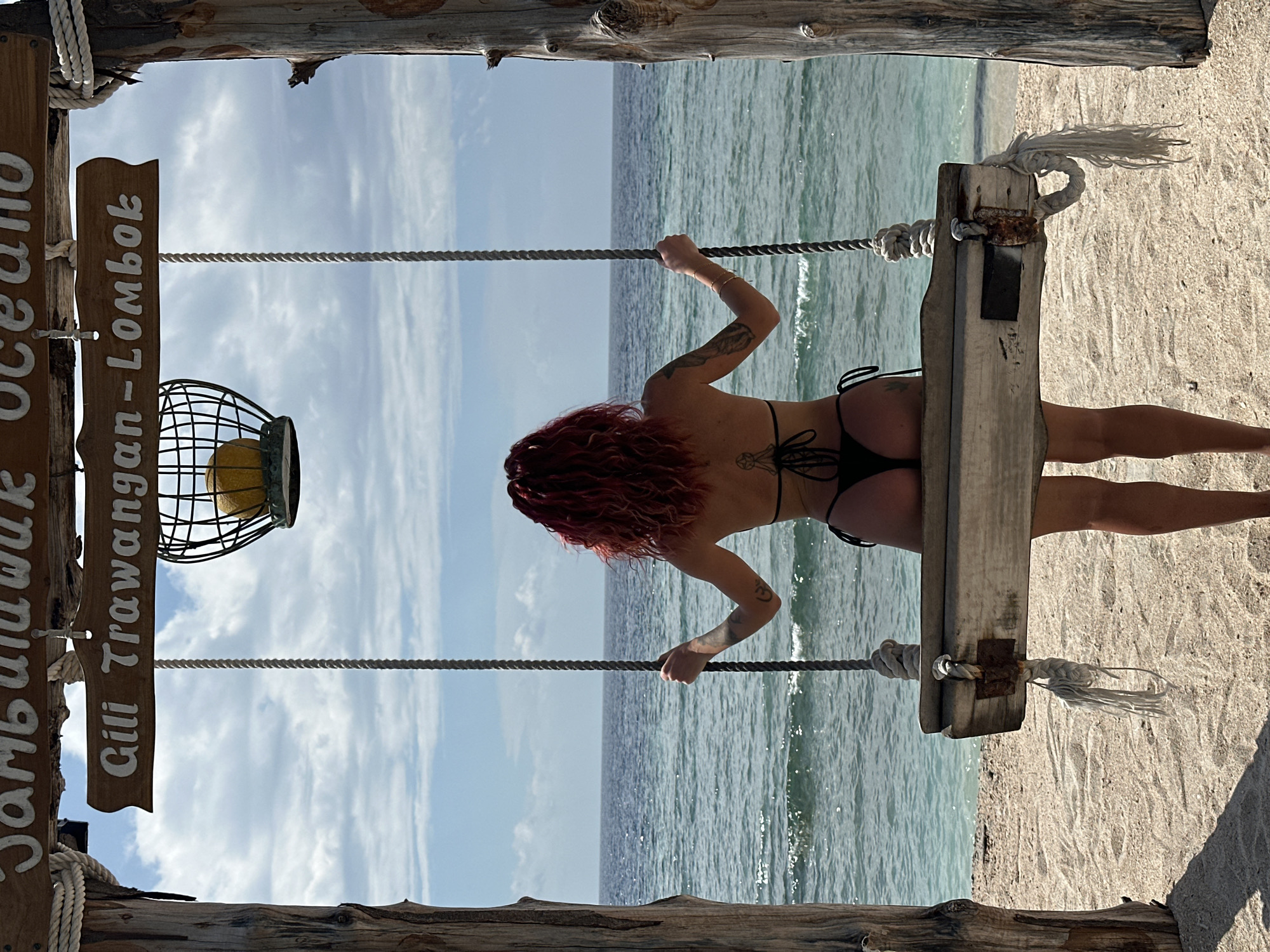 A vertical shot depicts a woman with red hair wearing a black bikini facing away from the camera, standing on a wooden swing on a beach. The swing is suspended by thick ropes connected to a wooden frame, and a sign with "Jambuluwuk Oceano Gili Trawangan - Lombok" is affixed to the structure. The background features the ocean under a sky with scattered clouds. The location is Gili Trawangan, Lombok, Indonesia.