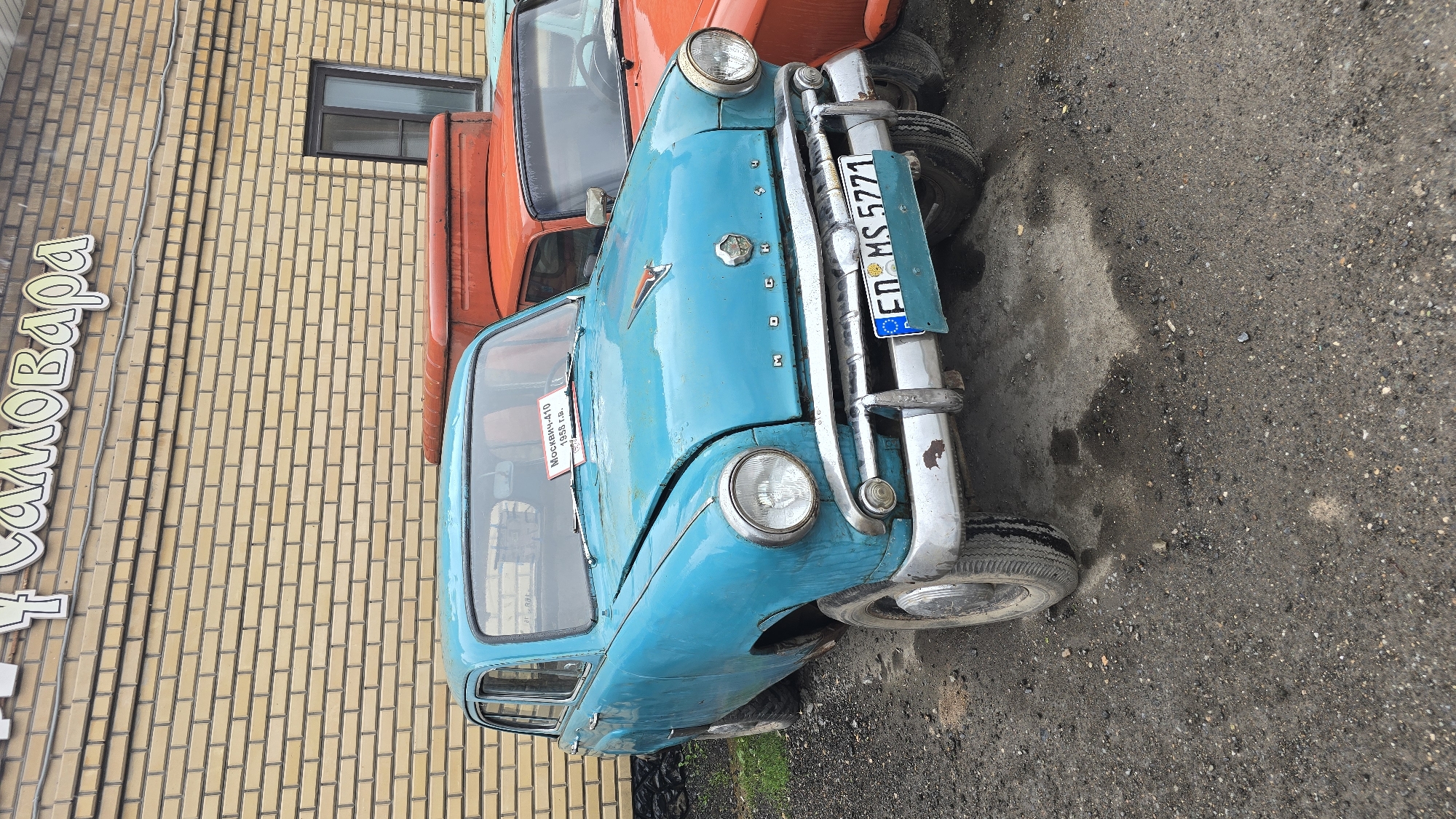 In Bol'shoi Mashlyak, Russia, a turquoise vintage Moskvich-410 car from 1958 is parked outdoors on a gravel lot in front of a light brown brick building. The building has a sign with Cyrillic text that reads "САМОВАРА" and a dark-framed window. The car has a chrome bumper, circular headlights, and a hood ornament. A paper sign on the windshield reads "Москвич-410 1958 г.в." and the license plate is "ED MS 5771". Another orange vehicle is partially visible behind the Moskvich. The ground is covered in gravel and there are a few dark wet spots, possibly from recent rain.