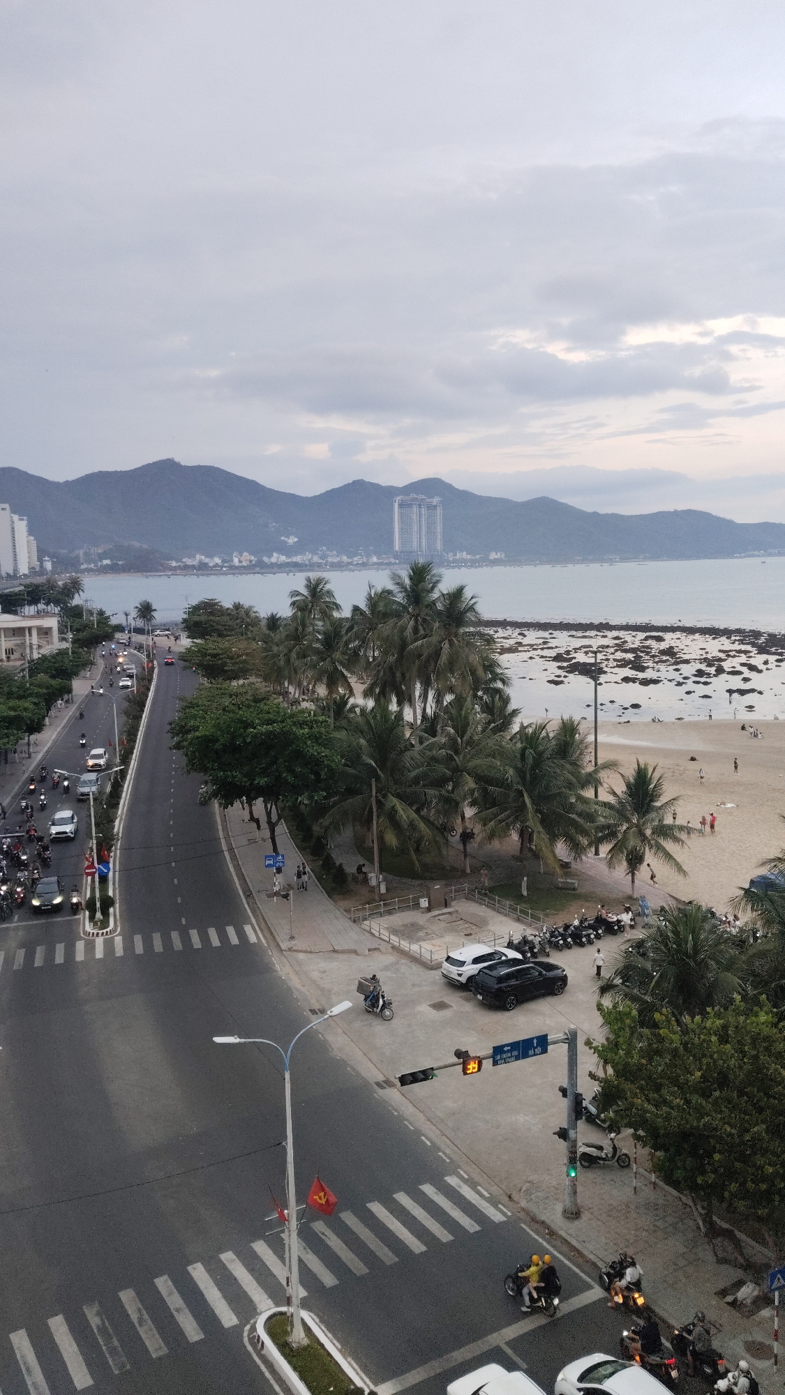 The image depicts a coastal scene in Nha Trang, Vietnam, specifically the Bac Nha Trang Ward. In the background are mountains with a tall building among them. The sea meets a sandy beach with scattered rocks and people engaging in various activities. Palm trees line the beach side of the street. The street itself is a multi-lane road with vehicular traffic including cars and scooters. A crosswalk is in the foreground with traffic lights showing a green light. Several vehicles are parked near the sidewalk, with pedestrians walking around. A Vietnamese flag is on a pole near the crosswalk. The sky is overcast.