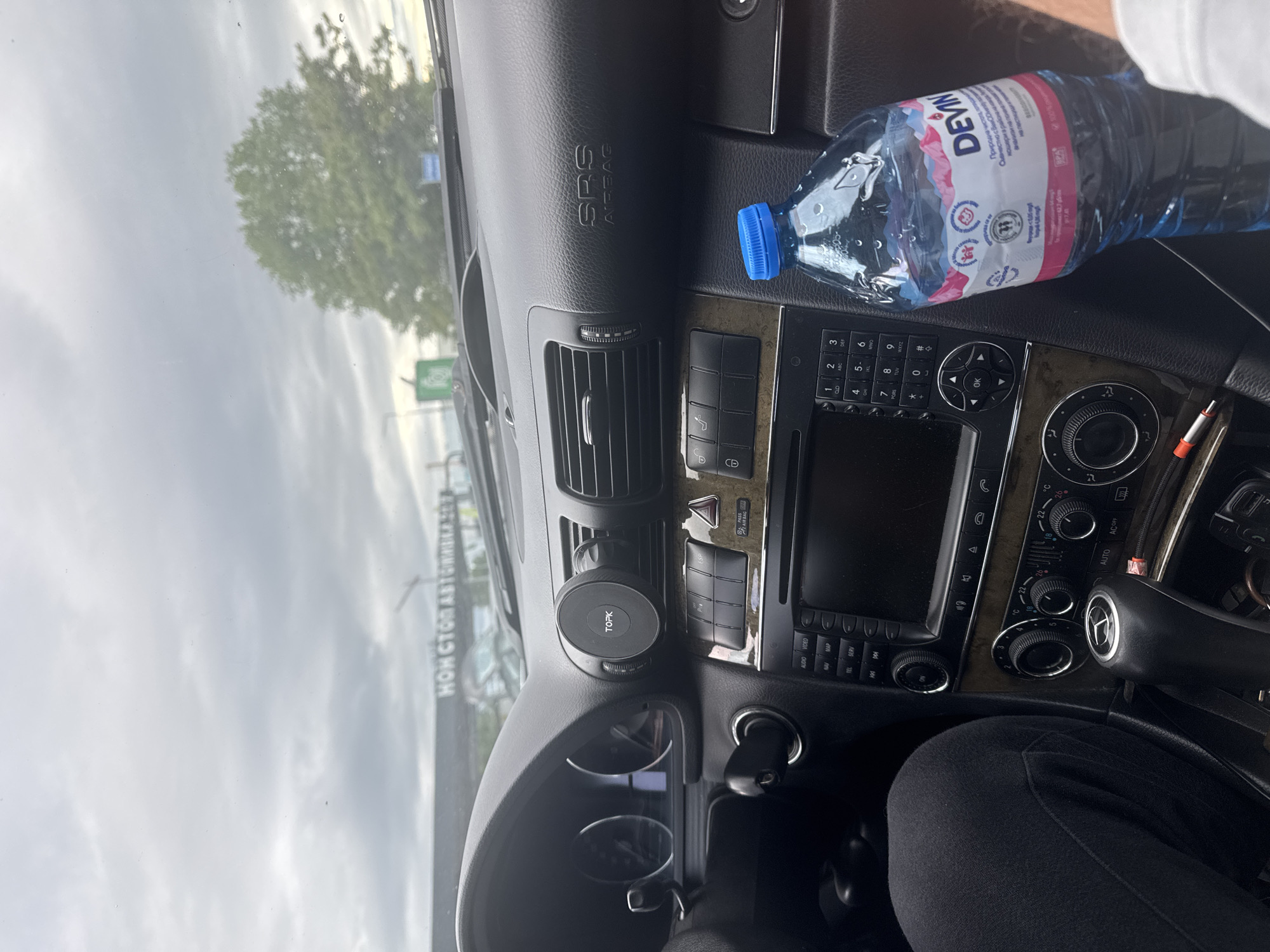 The image shows the interior of a car, specifically the dashboard. The dashboard is black with wood trim, and the SRS airbag indicator is visible. There is a water bottle with the label "DEVIN" lying on top of the dashboard. A "TOPK" branded phone holder is attached to the dashboard near the steering wheel. The car's radio and climate control panel are also visible.
Through the windshield, part of the outside environment is seen, including trees and a building with the text "НОНСТОПАВТGRADA.CO". Given the text "НОНСТОПАВТGRADA.CO" is a car service company with a location in Plovdiv, Bulgaria, it can be inferred that this is the city where the picture was taken.