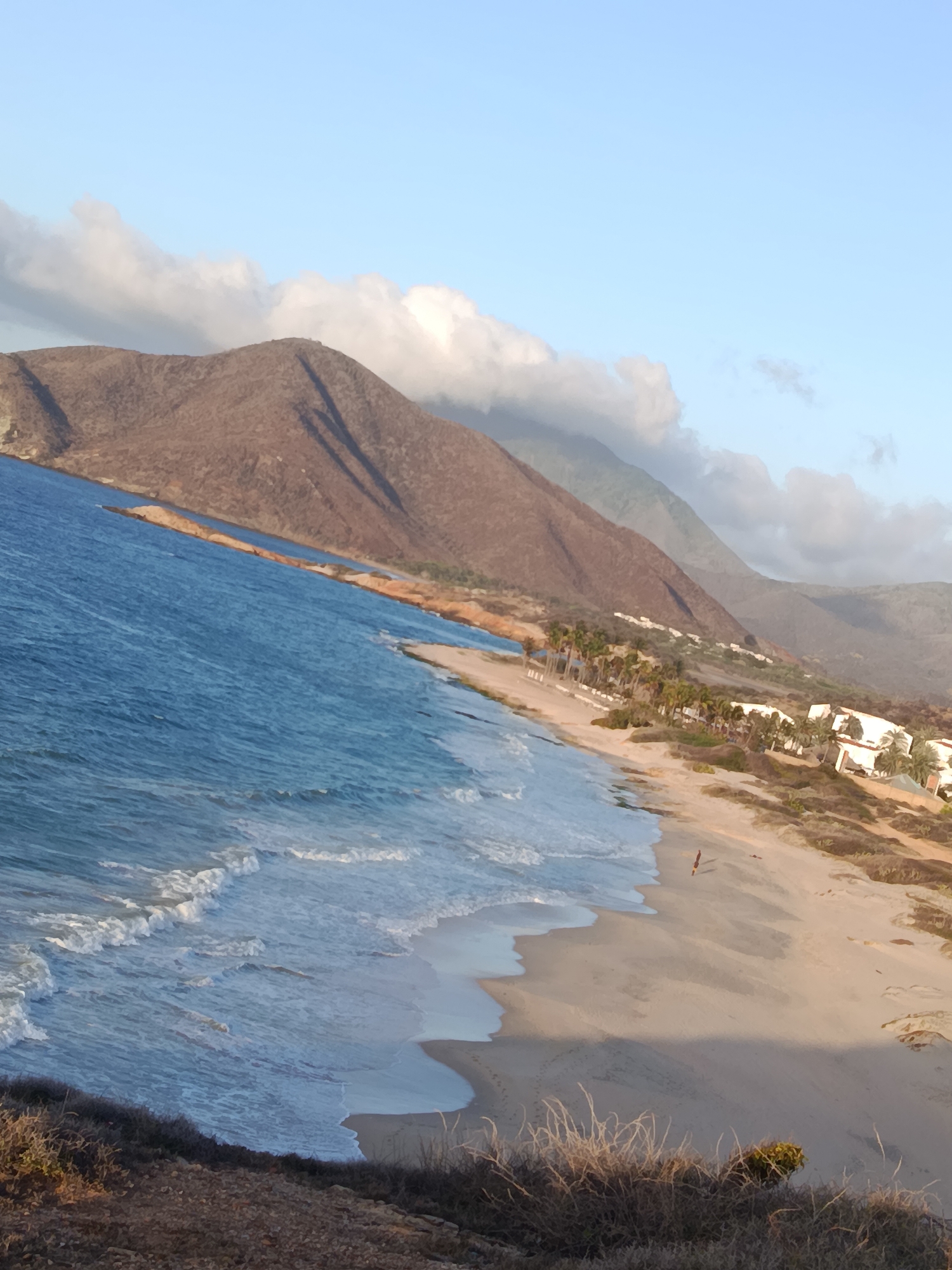The image shows a tilted view of a beach with gentle waves lapping at the shore. In the background, mountains rise up, with some buildings and palm trees visible along the coastline at the base of the mountains. The sky is a light blue with some scattered clouds. A lone person is visible walking on the beach. This scene is located in Altagracia, Venezuela, specifically at Playa Parguito, a well-known beach on Margarita Island. The lighting suggests it might be late afternoon or early morning.