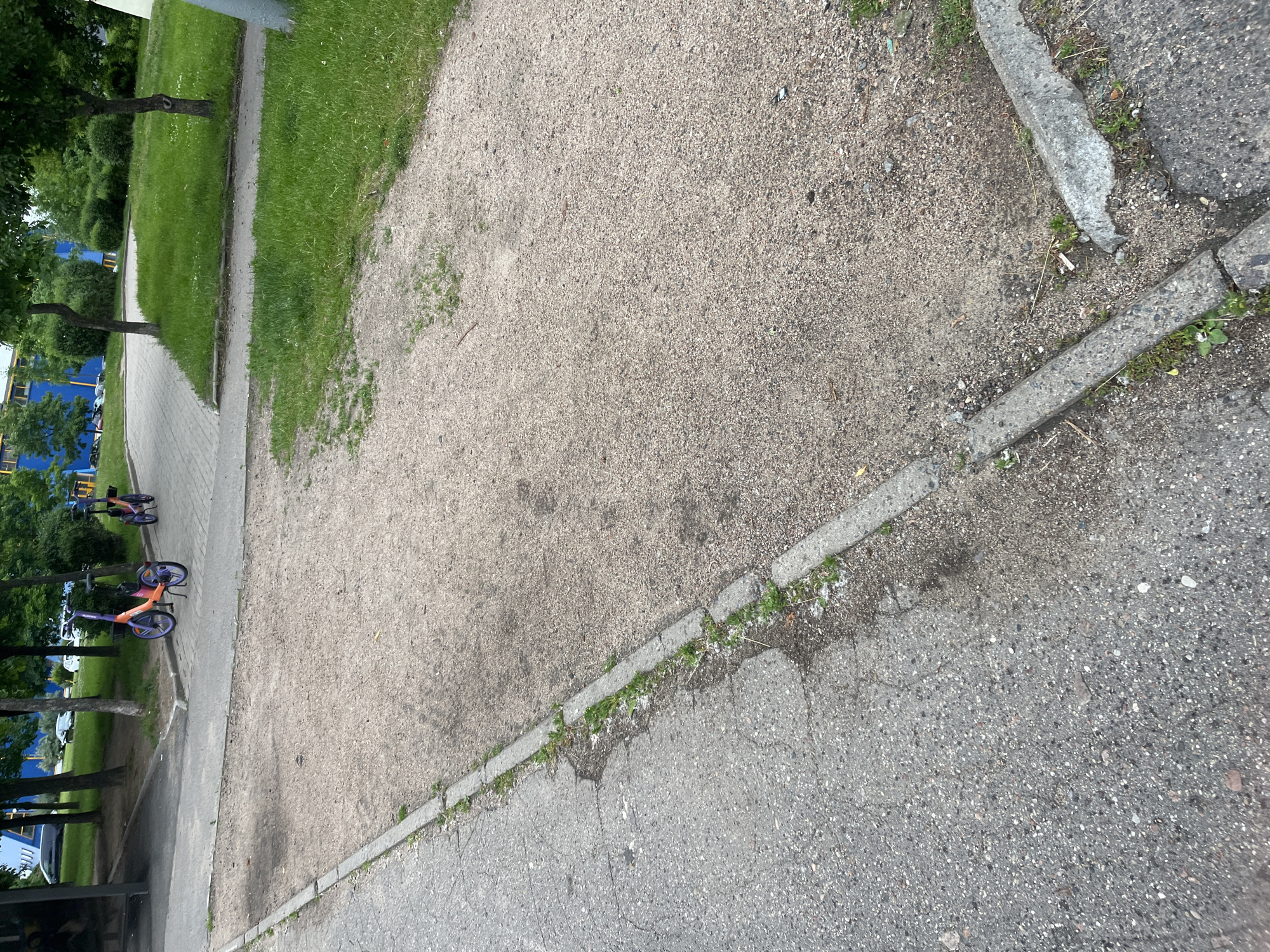 The image shows a sparsely populated urban outdoor environment, likely a public area in Minsk, Belarus. The scene features a paved walkway with two purple and orange electric scooters parked on it. The walkway is adjacent to a gravel path and a grassy area with trees. In the background, there are buildings, some with blue accents, and parked cars. The overall impression is one of a utilitarian public space, possibly a park or a designated parking area for shared mobility devices. The lighting suggests it is daytime, with a neutral overcast sky.
