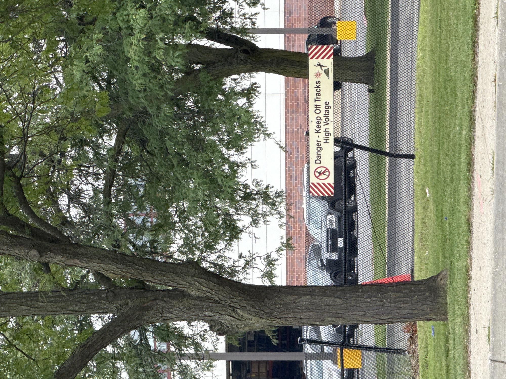 In Skokie, United States of America, the image captures an outdoor scene behind a black chain-link fence. Two large trees with rough bark and dense green foliage dominate the foreground. Behind the fence, several dark-colored trucks are parked. A horizontal yellow sign with red and white striped borders is attached to the fence, displaying text that reads "Danger - Keep Off Tracks High Voltage" along with warning symbols of a person and lightning. The background features a modern brick and white building with large windows. Green grass covers the ground in front of and behind the fence.