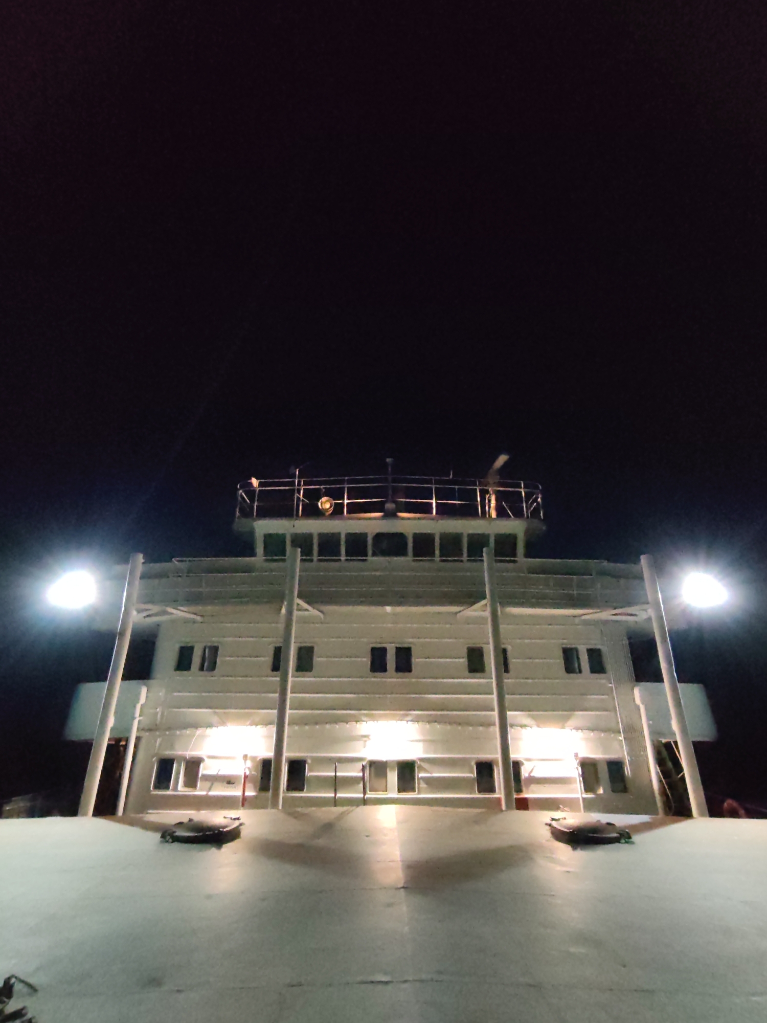 The image depicts the front of a ship at night. The ship is illuminated by lights along its exterior. The sky is dark, suggesting nighttime. The perspective is from the front of the ship, looking upwards towards the bridge. There are no people visible. There is no evidence in the image to confirm it is in Bol'shie Mkhi, Russia.