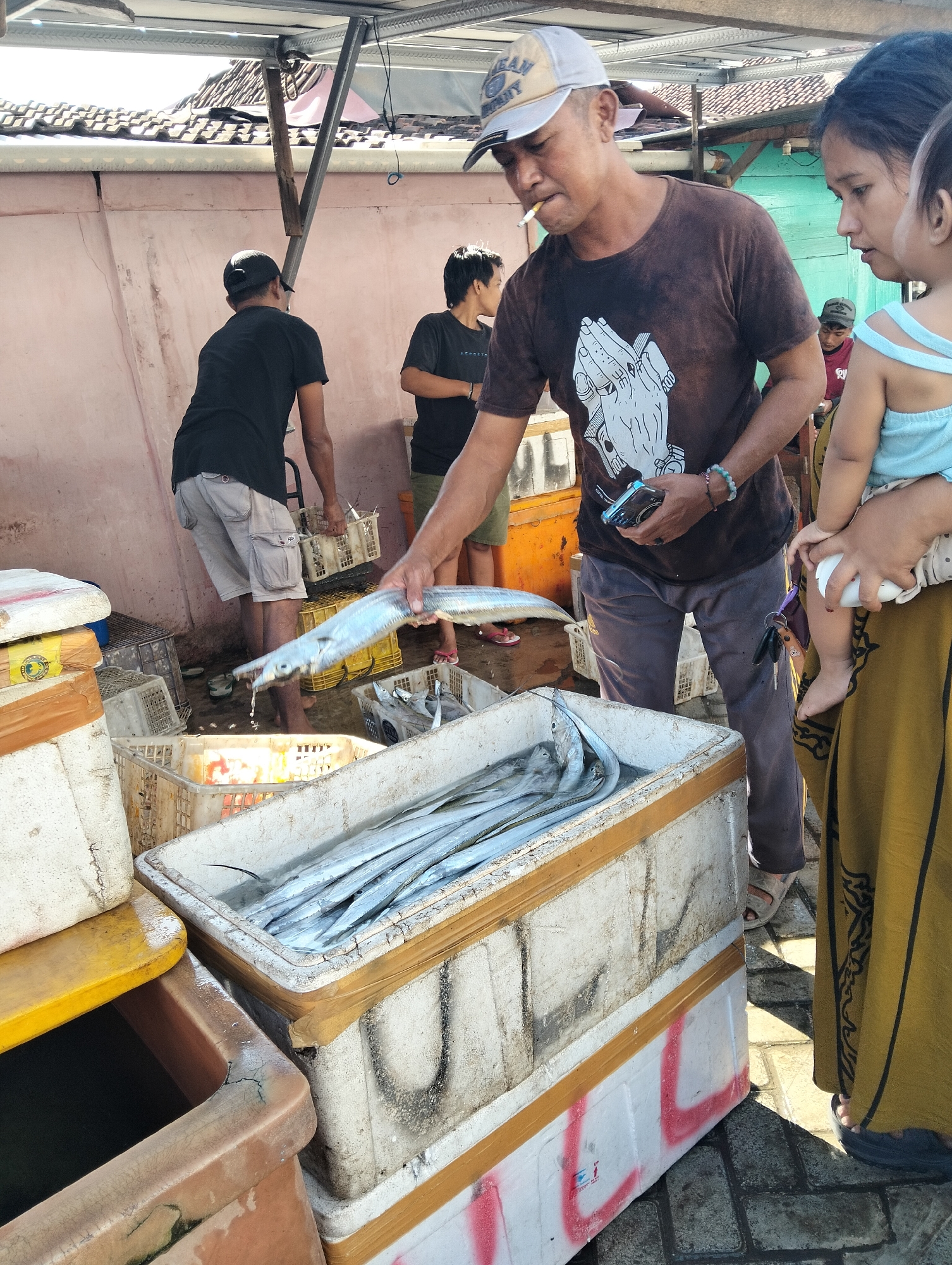In the city of Muncar, Indonesia, at a fish market, a man in a brown t-shirt and grey pants is holding a long, silvery fish above a white crate overflowing with similar fish. He is wearing a baseball cap and smoking a cigarette. To his right, a woman in a yellow dress holds a baby in her arms. Behind the man, another man in a black shirt and grey shorts is visible, and further back, a young person in a black shirt stands near a stall. The environment is outdoors, with a tiled roof structure overhead and a pink wall in the background. Various plastic crates and containers, some holding fish, are arranged around the scene. The ground is paved with bricks.