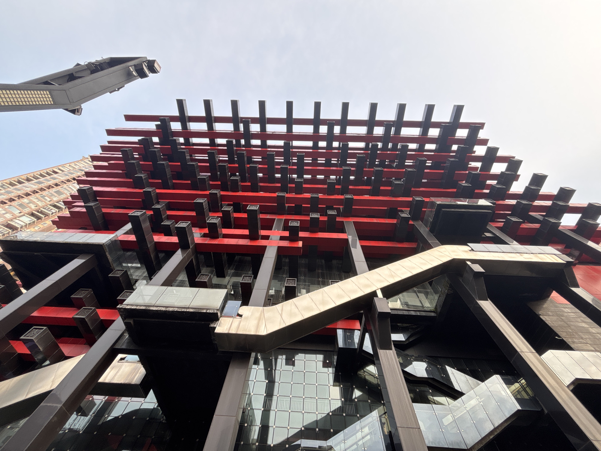 The photograph shows a low-angle view of the Raffles City Chongqing complex in Yuzhong District, China. The architecture features a network of red horizontal beams and black vertical supports, creating a grid-like pattern on the building's facade. Smaller black rectangular elements are interspersed throughout the structure. Glass windows and panels are visible between the beams. A diagonal, gold-colored structural element is prominent in the foreground. A street light is visible in the upper left corner of the frame. The sky is overcast.