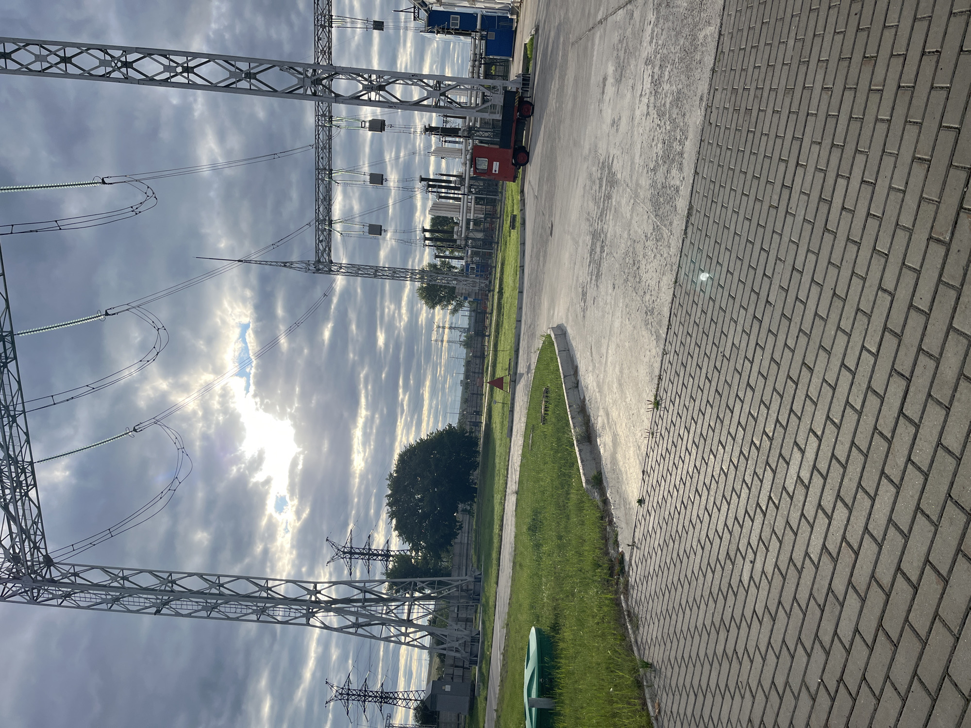 The image depicts an outdoor industrial area, likely part of an electrical substation, with a cloudy sky and sunlight breaking through. Dominating the scene are large metal lattice towers supporting high-voltage power lines. In the foreground, there's a paved walkway made of grey interlocking bricks, bordered by a concrete curb. Adjacent to the walkway is a grassy area with a green circular object, possibly a manhole cover or a safety barrier. A paved road or service area extends into the mid-ground, where a small red utility vehicle with three wheels is parked. Beyond the vehicle, there are various industrial structures, including electrical equipment, pipes, and buildings. In the background, more power line towers are visible, along with trees and a distant fence. The overall environment suggests an operational facility with infrastructure related to power transmission. This scene is located in Chaikovsky, Russia.
