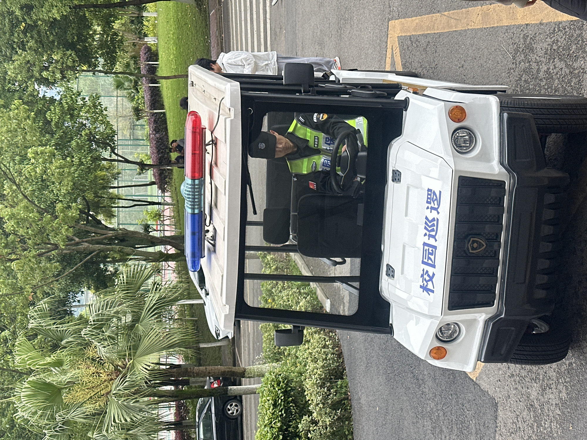 The image, taken in Jiangjin, China, displays an outdoor scene featuring a white utility vehicle and several individuals within a green, well-maintained environment, likely a university or school campus.
In the foreground, a white, open-sided patrol vehicle is parked on an asphalt road with faded yellow markings. The vehicle is equipped with a red and blue emergency light bar on its roof. Its front features headlights, amber turn signals, a dark grille, and blue Chinese characters that read "校园巡逻" (xiàoyuán xúnluó), meaning "Campus Patrol."
A person, presumably a security guard, is seated in the driver's seat. They are wearing a dark cap, a dark uniform shirt, and a conspicuous yellow-green reflective vest with additional Chinese text, partially visible, related to security. Their hands are on the steering wheel, and their gaze is directed forward.
In the background, behind and to the side of the vehicle, a lush green lawn is visible, dotted with various trees, including prominent palm trees and other deciduous types, along with bushes and hedges. Several other people are present in this area; one individual stands near the rear of the patrol vehicle, dressed in a white shirt and grey pants, while others are seen sitting or standing casually among the greenery, suggesting a relaxed atmosphere. A fence can be discerned further in the distance, indicating a boundary or a sports facility.
The scene is brightly lit by natural daylight, consistent with daytime conditions. The light is soft and even, implying an overcast sky or diffused sunlight. The overall setting conveys a sense of routine activity on a well-maintained campus.