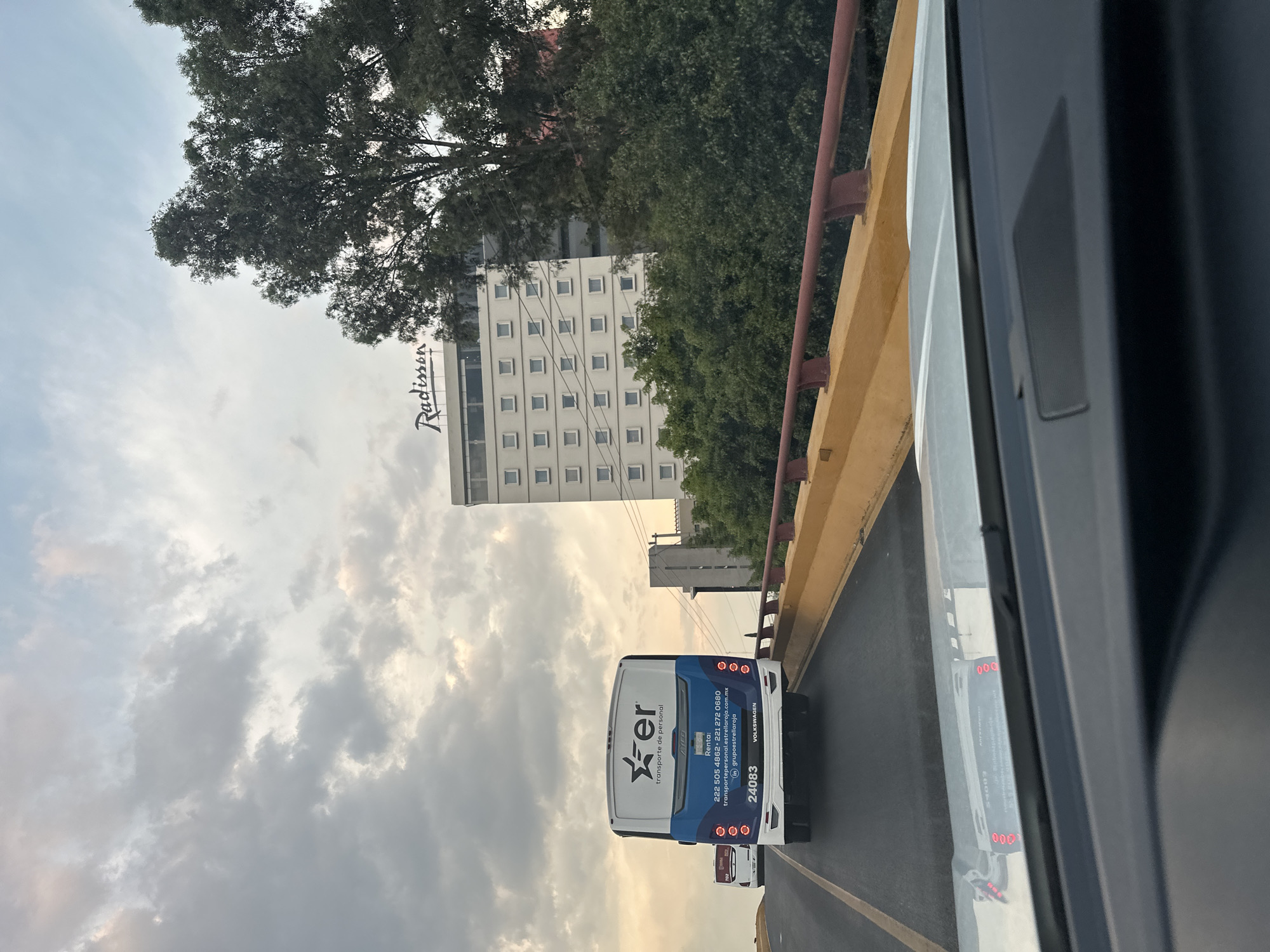 The image is taken from the inside of a vehicle, potentially a bus or car, and captures a street scene in Puebla, Mexico. A Radisson Hotel is prominently displayed to the left, with the hotel's sign clearly visible. Adjacent to the hotel is a tall tree that partially obscures the building. In the foreground, there is a highway overpass with a mustard yellow and red safety rail. The photograph also shows an "er" bus with the text "transporte de personal" and the phone number "222 505 4862-221 272 0680." The bus also displays the license plate number "24083" and the brand name "VOLKSWAGEN". The sky is cloudy.