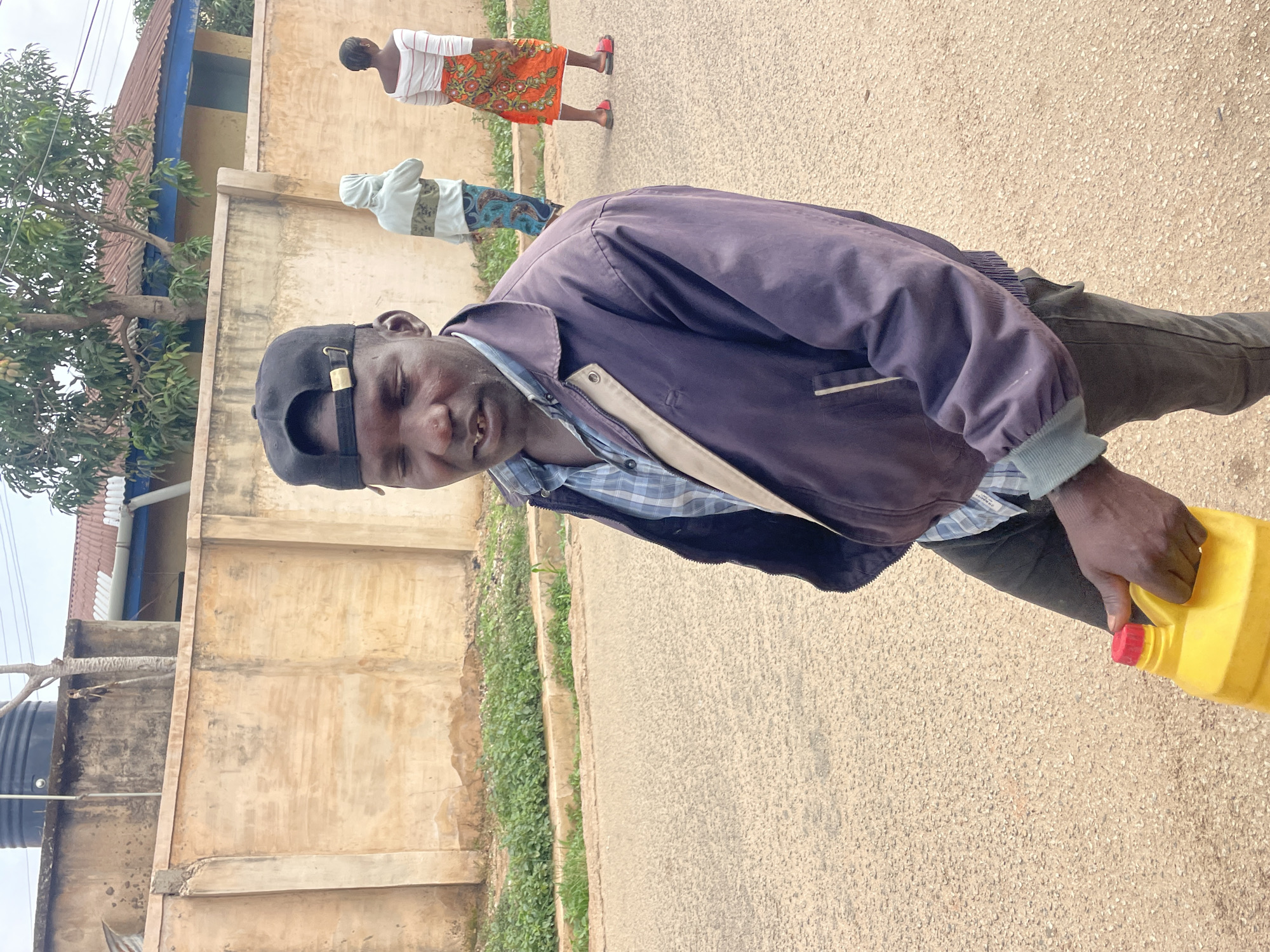 A man wearing a dark-blue jacket and a black cap is walking on a road. He is holding a yellow container with a red cap. In the background, two people are walking. The background includes buildings with visible walls and a water tank. The location appears to be a street in Jos, Nigeria.