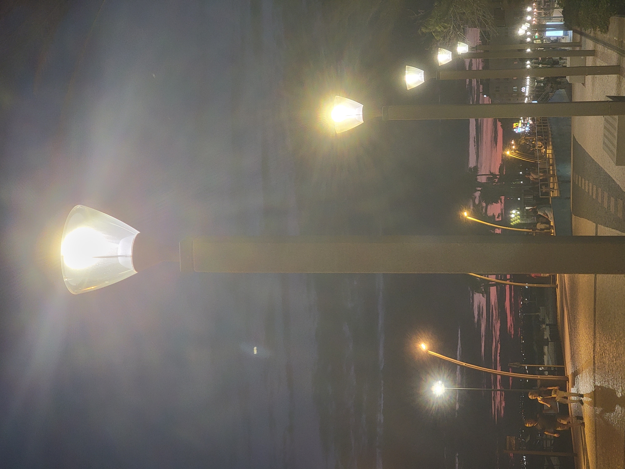 The image depicts a waterfront promenade in Quarteira, Portugal, at dusk. The sky is a deep indigo with streaks of pink and orange from the setting sun, suggesting a warm evening. Multiple streetlights with translucent conical shades illuminate the walkway. In the foreground, two individuals, a man and a woman, are walking side-by-side on the paved path. The man is wearing a dark shirt and pants, and the woman is wearing a light-colored top and pants with a crossbody bag. Their reflections are visible on the wet pavement, indicating recent rain or cleaning. Further down the promenade, more streetlights and distant city lights are visible, creating a festive atmosphere. Trees and architectural elements line the promenade, and a low blue wall separates the walkway from a lower level. The overall scene conveys a peaceful and inviting ambiance characteristic of a coastal town at twilight.