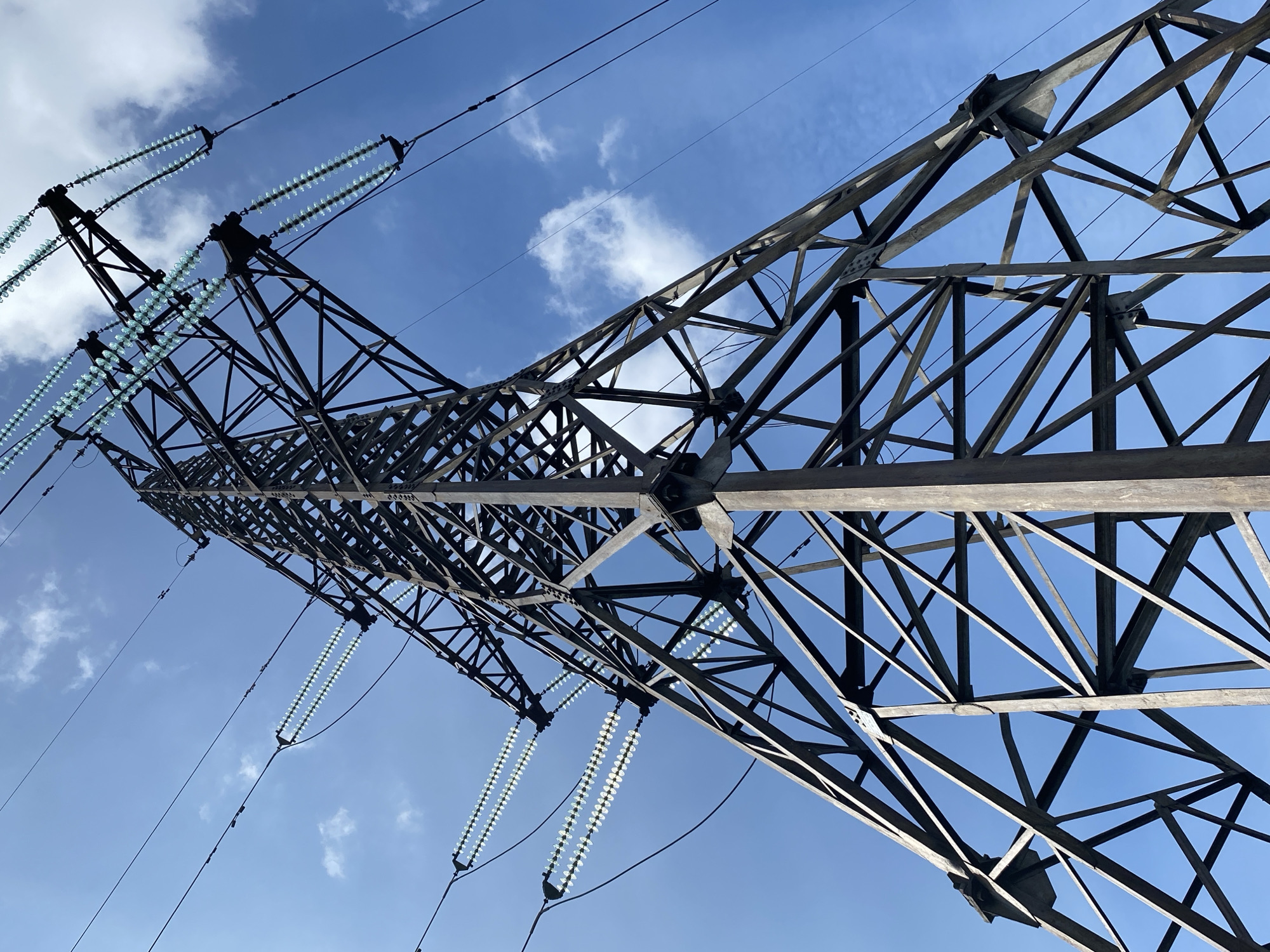 The image shows a low-angle view of a high-voltage power line tower against a blue sky with scattered clouds. The tower is constructed of steel lattice beams. Attached to the tower are insulators with numerous circular glass or ceramic discs strung together. Overhead power lines are connected to the insulators. The location is Chaikovsky, Russia, as indicated by the photographer and subject of the image. The scene presents an infrastructure feature of electricity transmission.