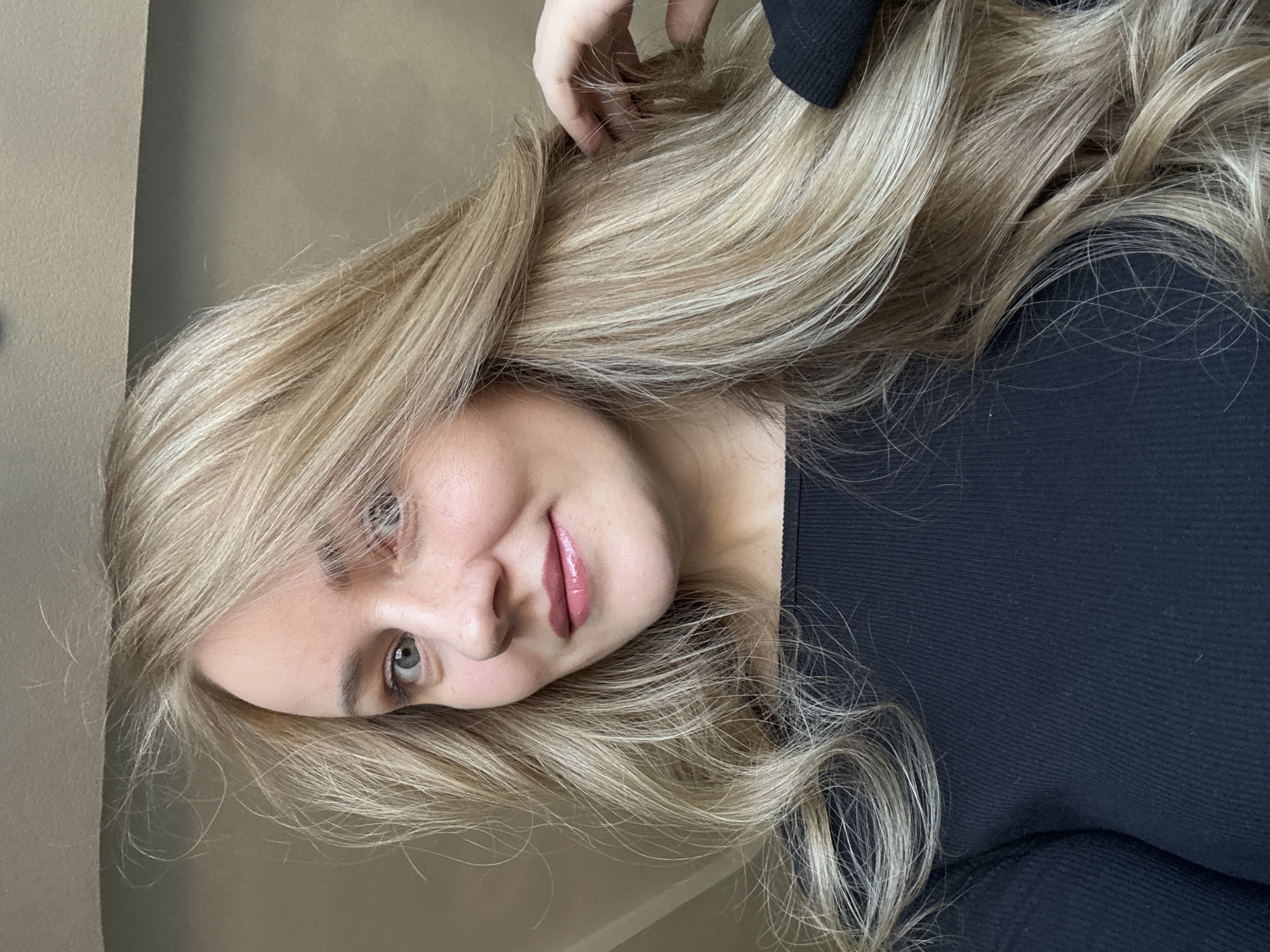 A fair-skinned woman with long, wavy blonde hair is angled in the center of the frame, wearing a black, ribbed long-sleeved shirt. She is looking at the camera with a slight smile, her hand touching her hair. The background is a neutral beige wall. The location is unknown.
