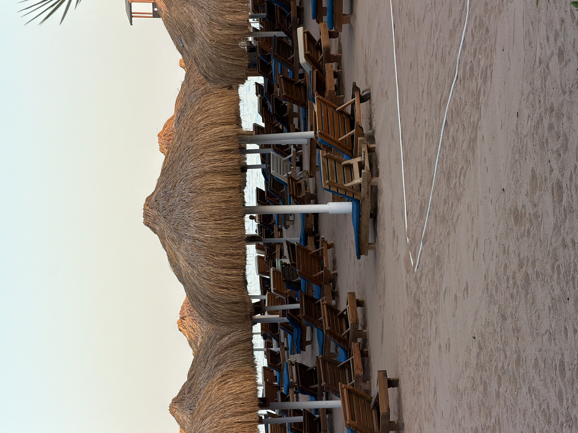 The image depicts an empty beach resort in Sharm El Sheikh, Egypt, with rows of unoccupied lounge chairs and thatched umbrellas lined up on the sand. The beach appears to be deserted, with no visible people in the immediate foreground or middle ground. In the background, there are glimpses of the sea and a faint silhouette of a person on a distant lounge chair. The sky is clear and pale, suggesting either early morning or late evening light. A white rope or cable is visible on the sand, dividing the beach area. The scene conveys a sense of tranquility and stillness.