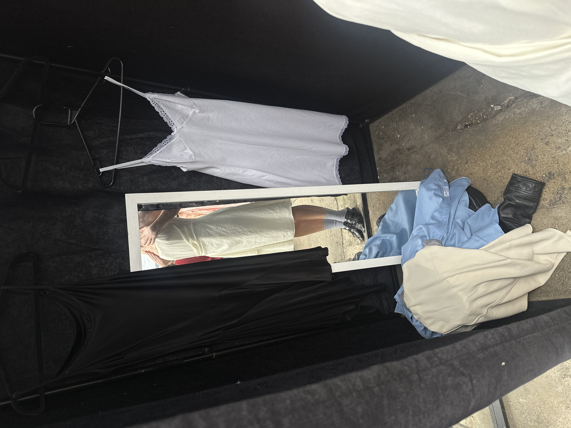 The image shows a collection of clothes and a mirror placed within what appears to be a makeshift outdoor display or installation. Garments include a white dress with lace trim on a hanger, a black dress on a hanger, and a pile of clothes consisting of a light blue shirt, a cream-colored shirt, and a black leather boot. A rectangular mirror reflects a person wearing a yellow dress, gray socks, and black shoes. The background is dark, possibly consisting of black fabric or walls forming an enclosure, with an area of concrete visible on the ground. The location appears to be outdoors, potentially in an alley or courtyard. The image does not indicate a location in the city of London.