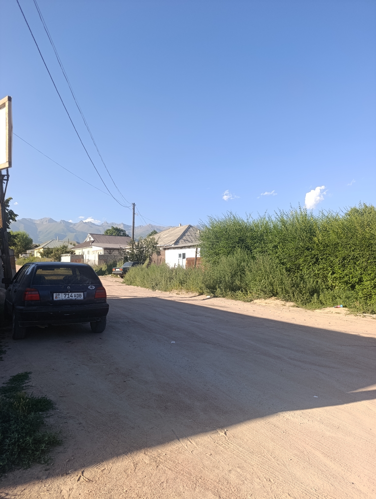 The image depicts a dusty, unpaved road in a rural setting, likely Bosteri, Kyrgyzstan, with houses and lush green trees lining one side. The sky is a clear, vibrant blue with a few wispy clouds. A dark-colored Volkswagen Golf is parked on the left side of the road, its rear facing the camera. The car's license plate is visible and reads "09 714 ADB". In the background, a line of mountains with rugged peaks stretches across the horizon. The scene appears to be captured on a sunny day.